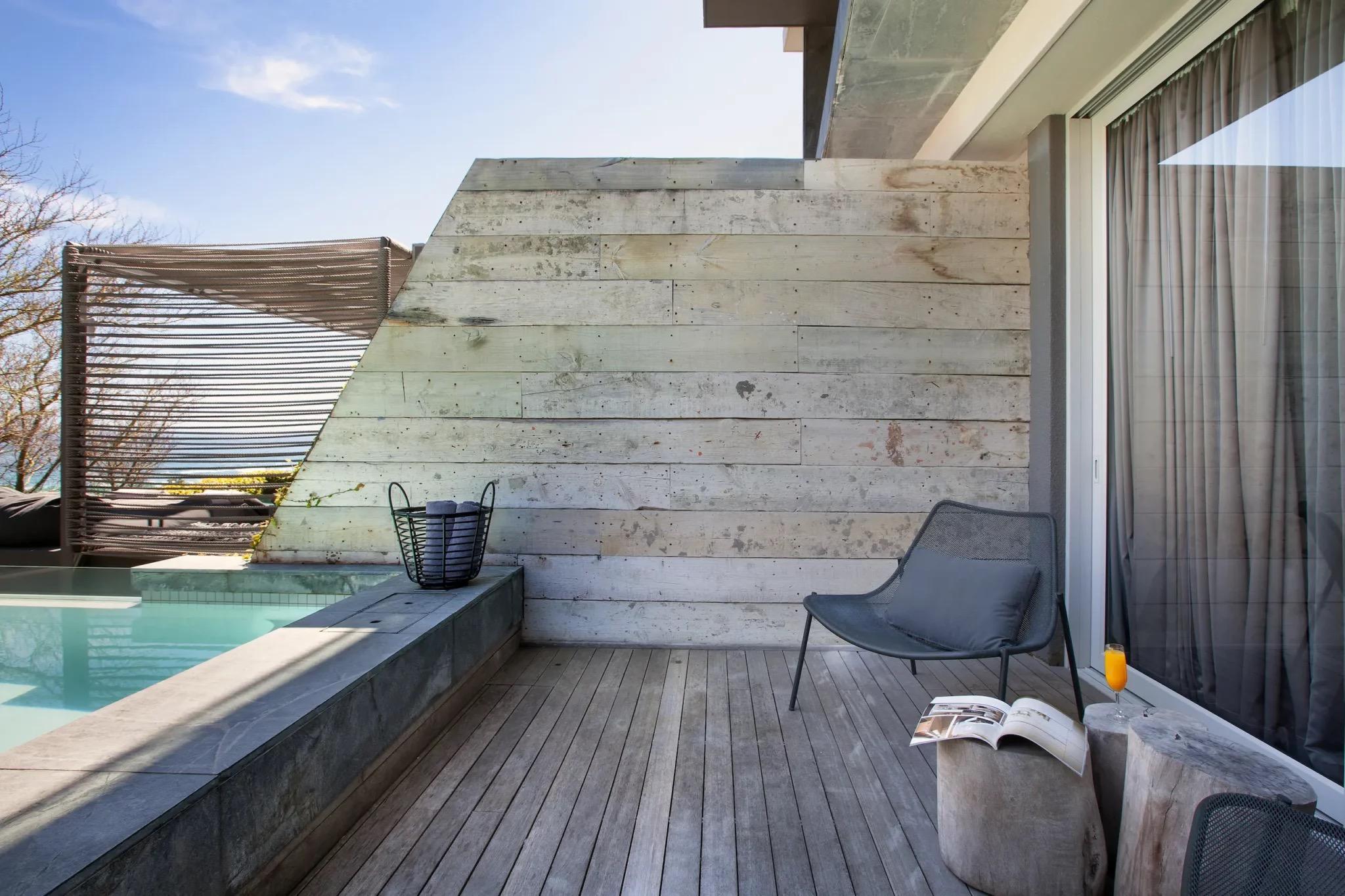 A private wood-decked terrace with a small plunge pool, a woven daybed, and a lounge chair with an open magazine and a drink on a tree-stump side table.