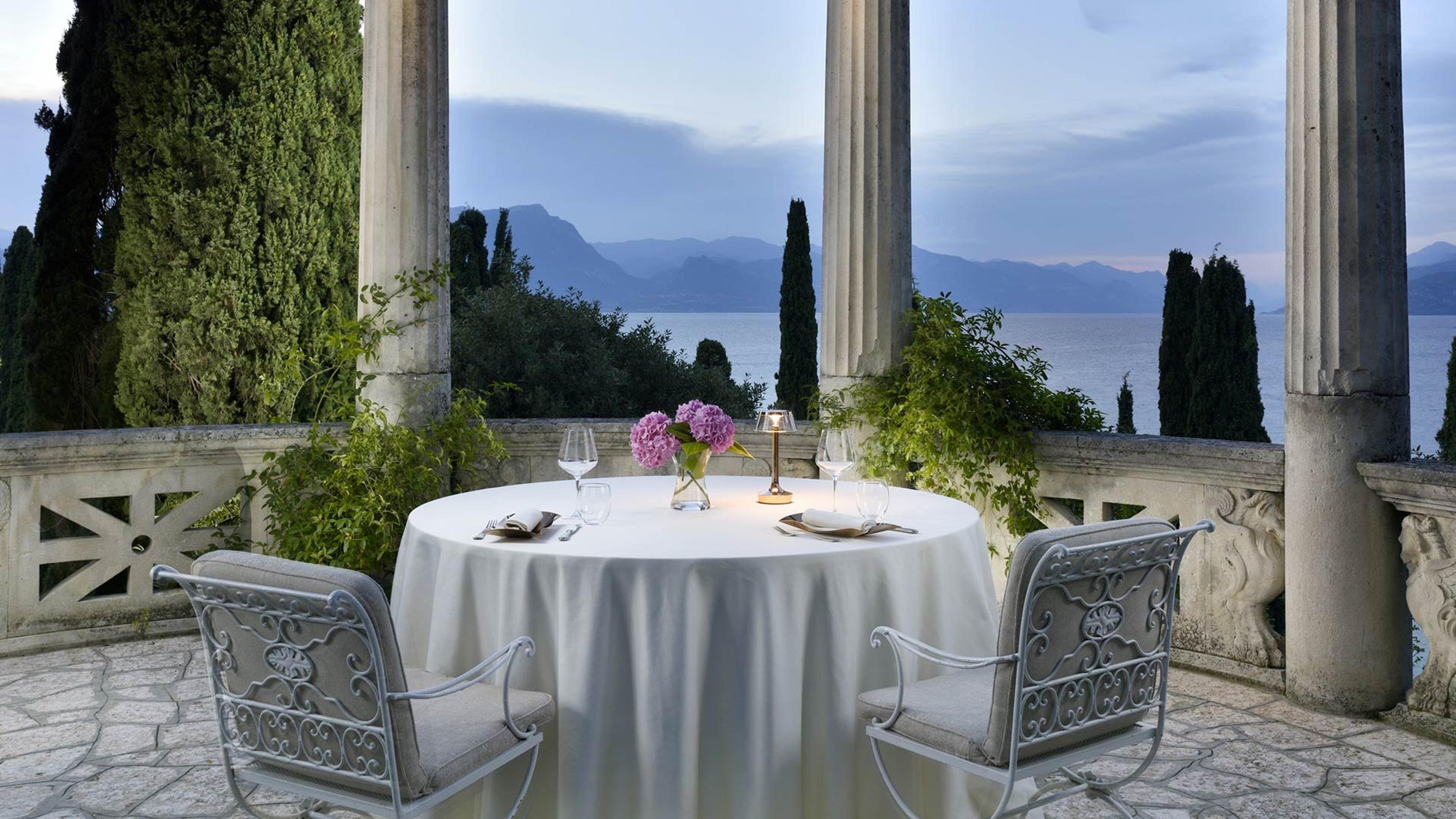 A round table set for two on a colonnaded terrace at dusk, with hydrangeas on the table and a lake and mountains beyond.