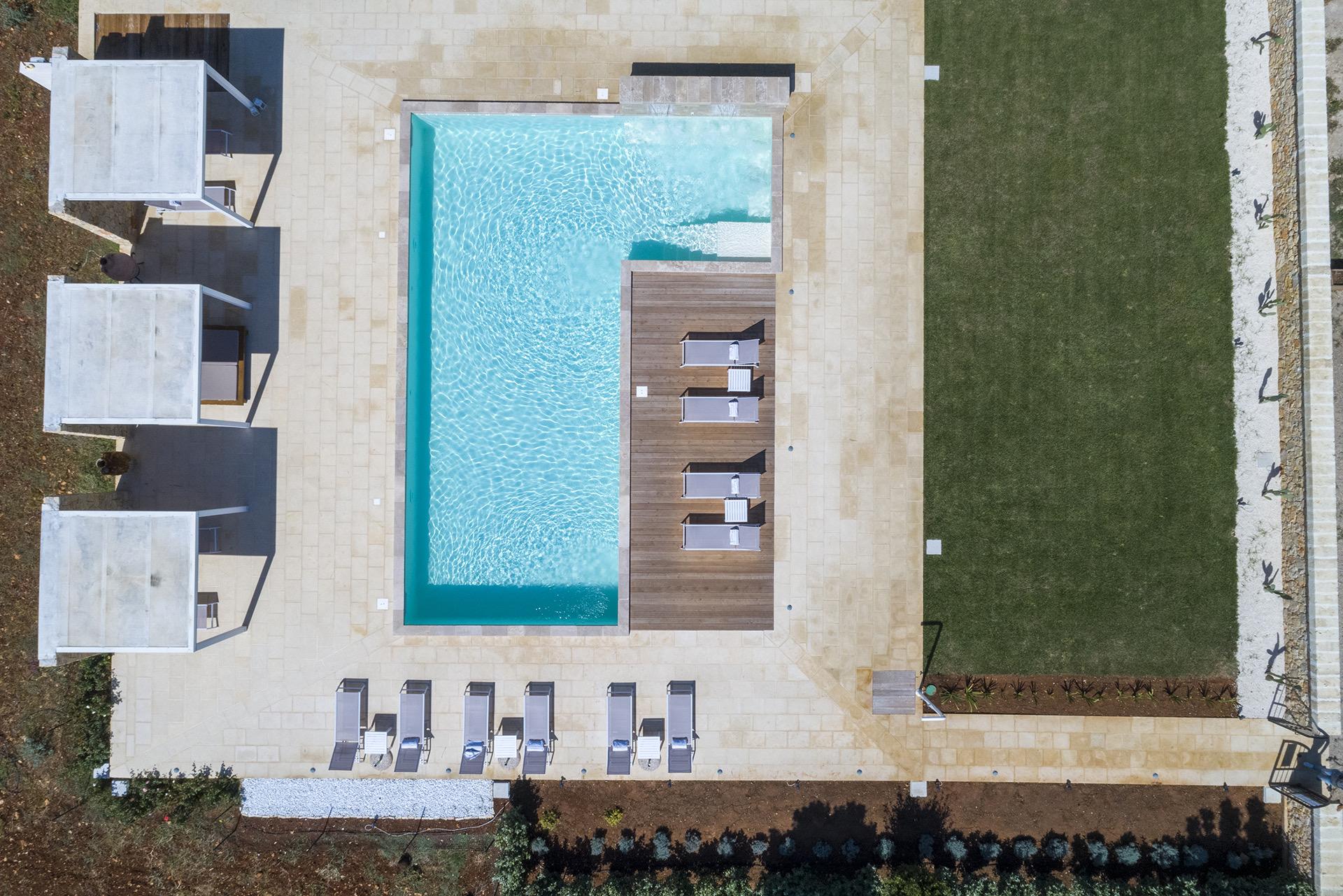 An aerial view of a pool with a sun deck, three stone cabanas and a row of sunbeds beside an open lawn.