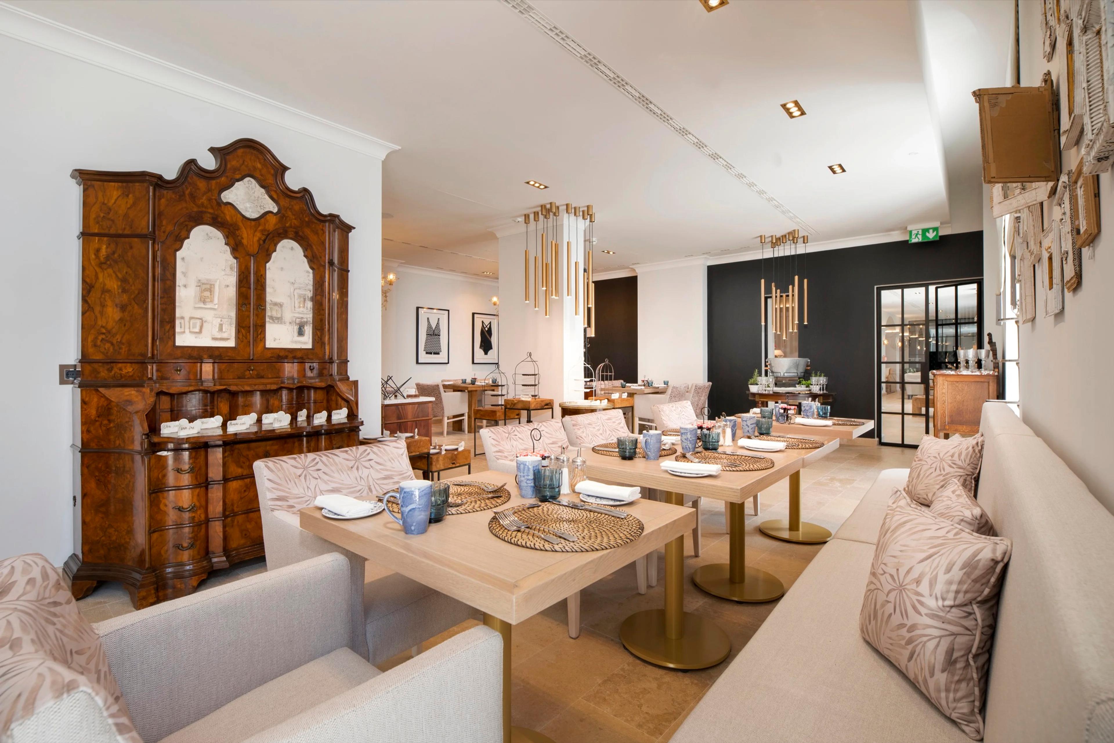 A restaurant dining room with a carved wooden cabinet, patterned armchairs, and small tables set with woven placemats and blue glassware.