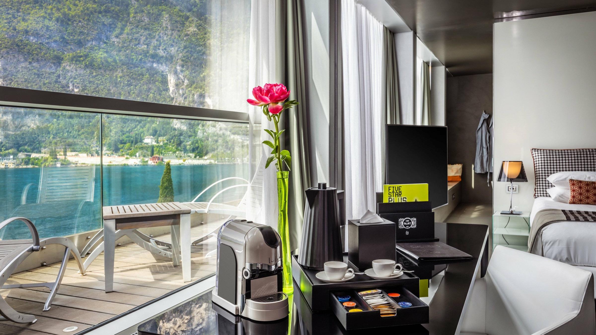 A hotel balcony with a coffee machine and a pink flower in a vase on a table, with sun loungers and a lake view beyond the glass railing.