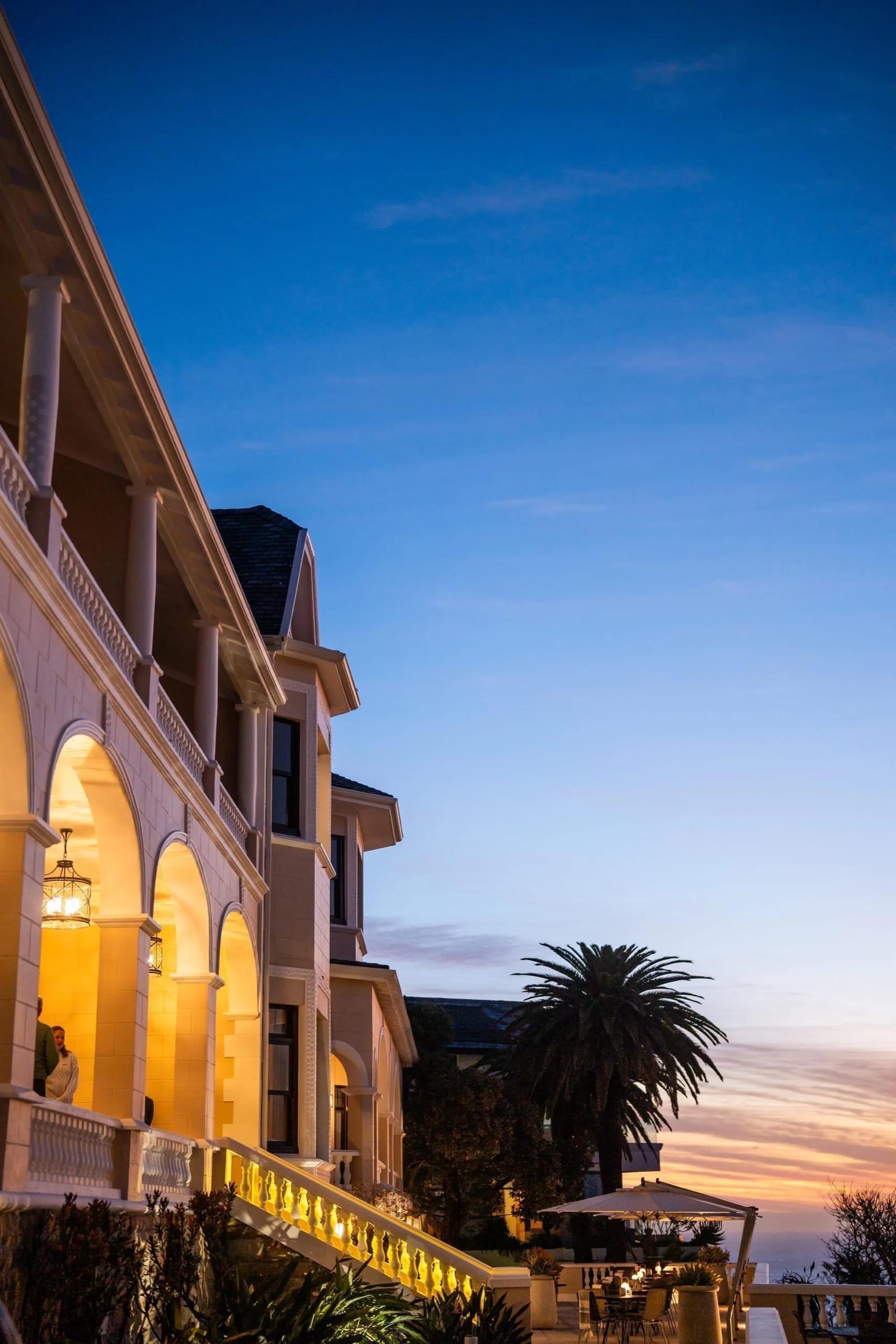 A two-storey cream building with arched colonnades and lit windows at dusk, with a palm tree and a parasol on the terrace below.