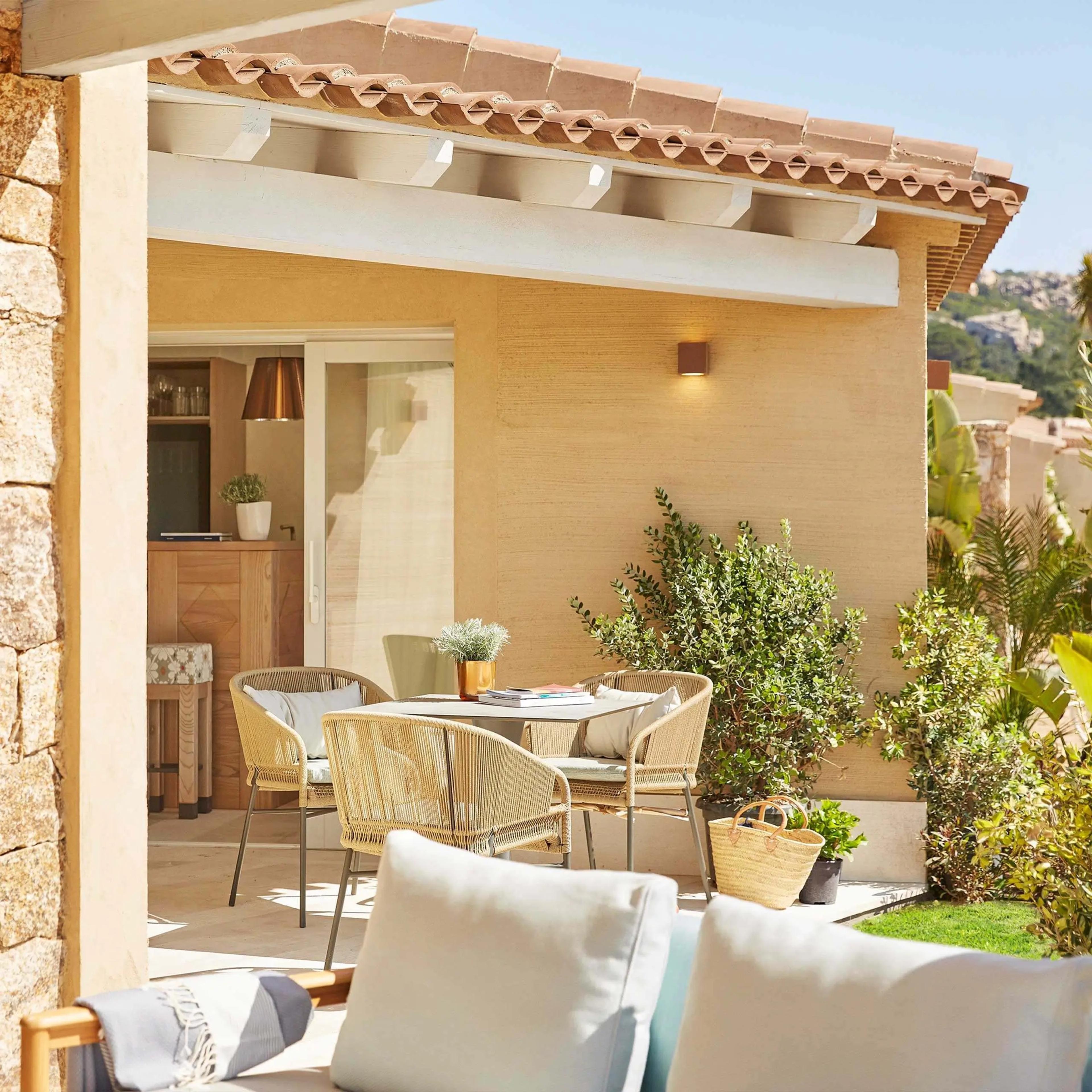 A shaded stone terrace with a rattan dining table and chairs outside a tiled roof building, Mediterranean shrubs along the wall.