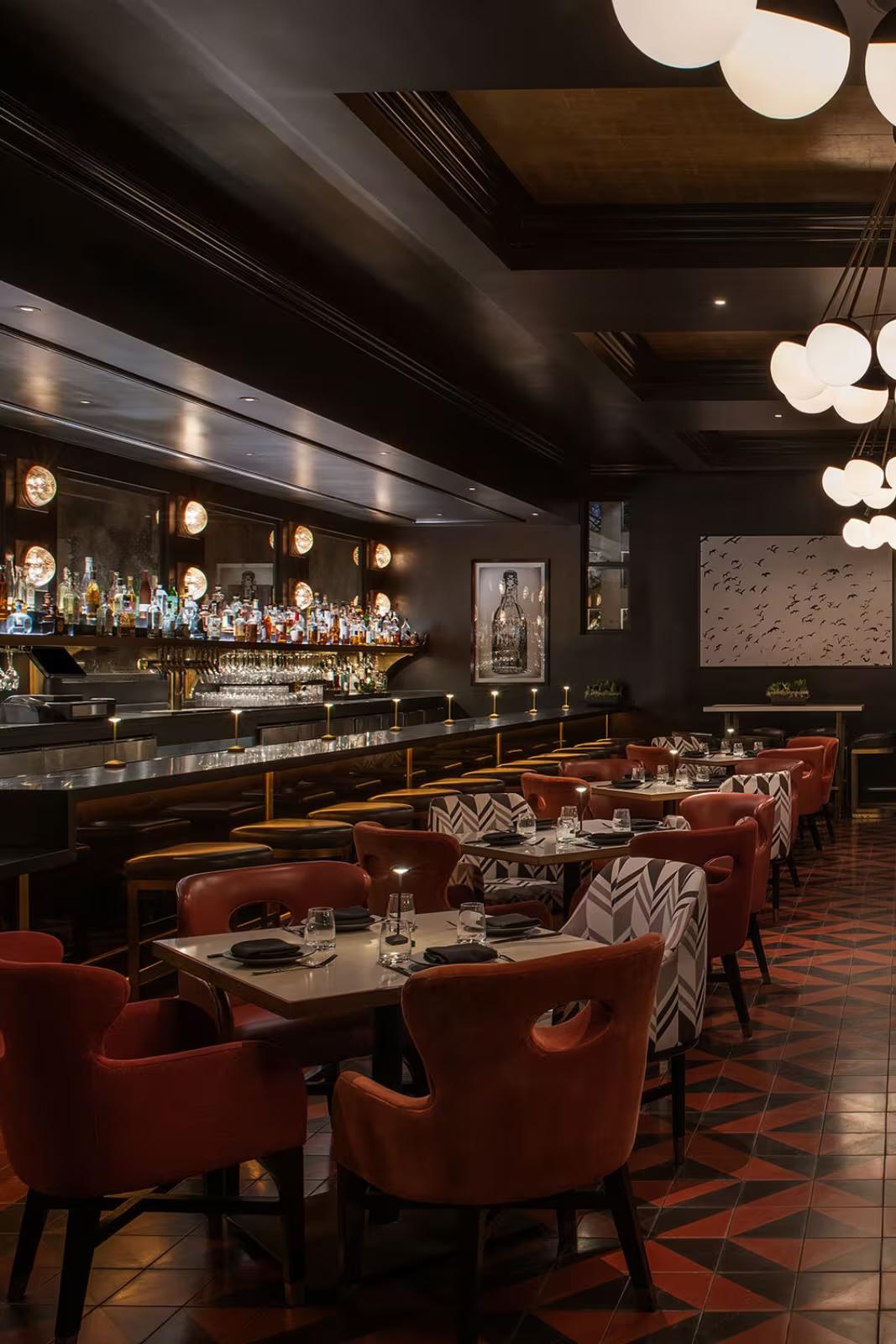 A dimly lit restaurant and bar with a long bottle-lined bar counter, orange upholstered chairs, patterned tablecloths, and globe pendant lights.