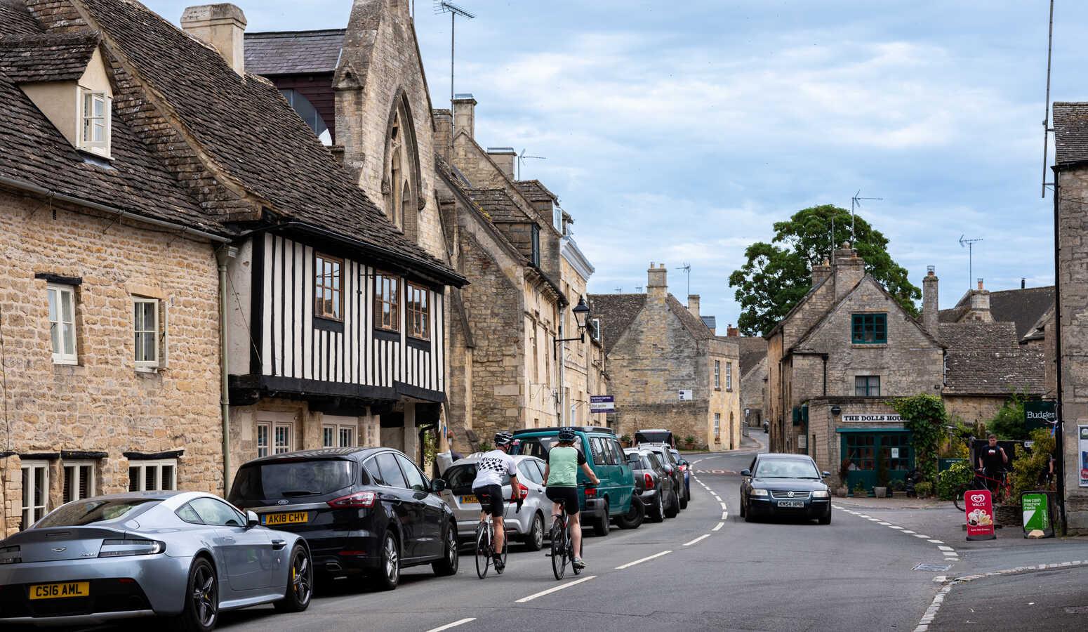 A Cotswold stone high street with terraced buildings, parked cars and two cyclists riding past, under a partly cloudy sky.