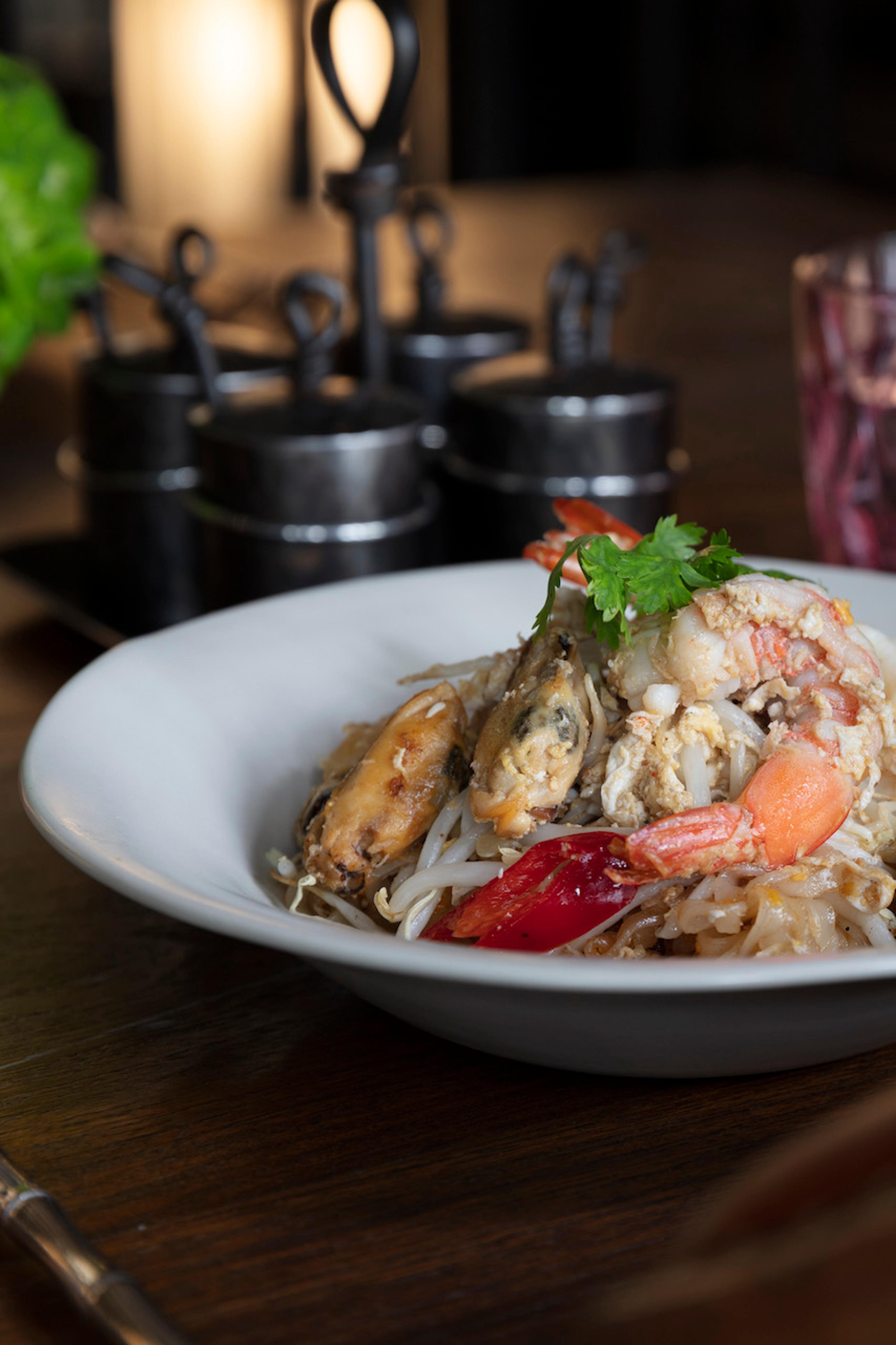 A white bowl of stir-fried rice noodles with prawns, mussels and bean sprouts sits on a dark wood table beside a glass and a row of small condiment jars.