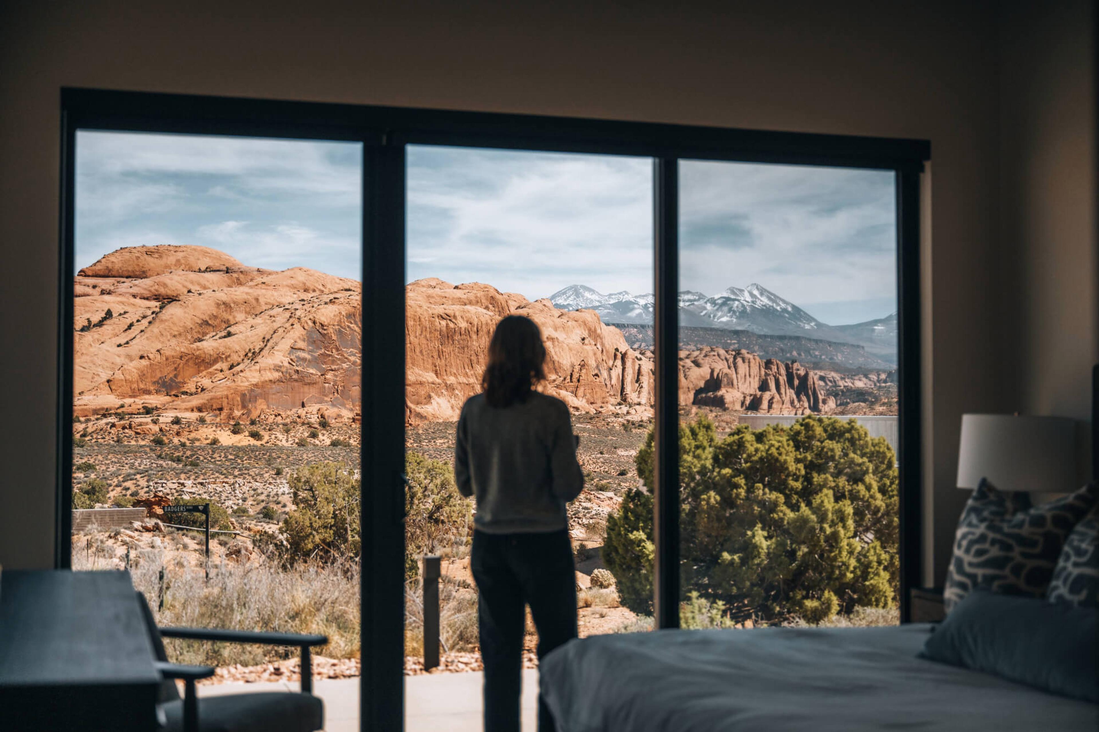 A woman seen from behind looks out a large window at red rock formations and mountains, standing in a bedroom with a visible bed and chair.