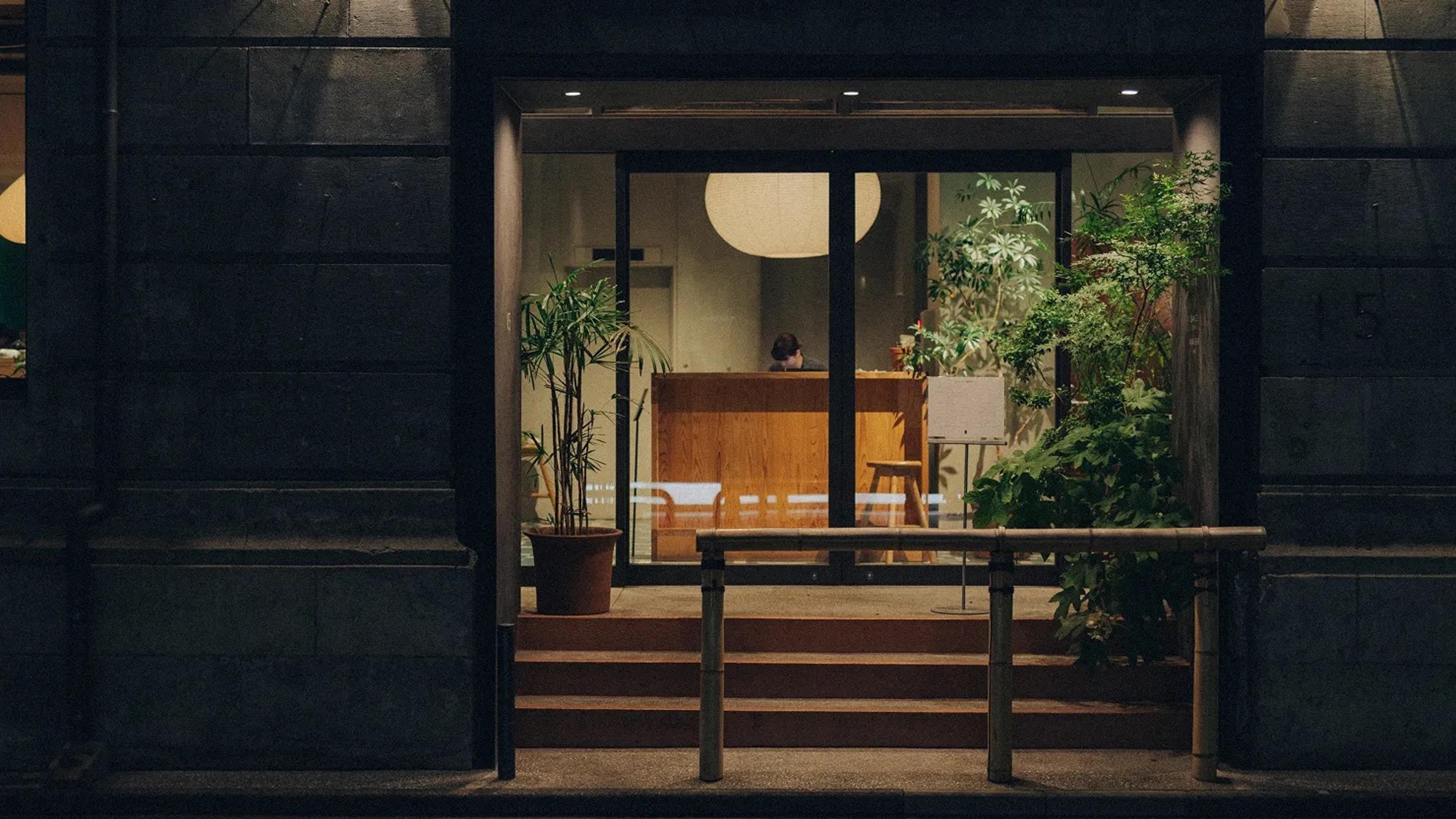 A lit glass entrance seen from the street at night, with a person seated at a wooden reception desk behind potted plants and a round paper lamp.