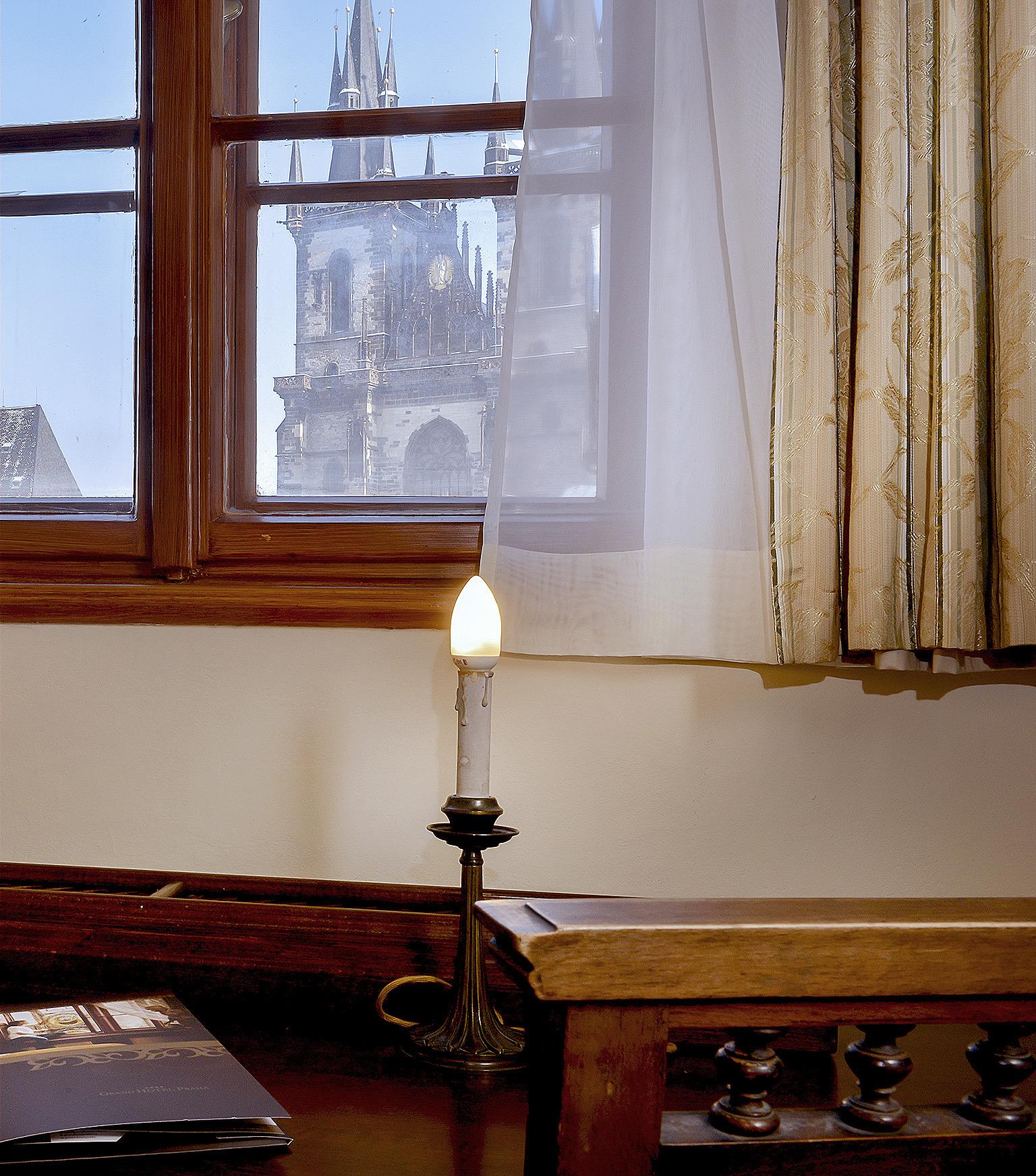 A lit candle on a windowsill in front of a sheer curtain, with gothic church spires visible outside.