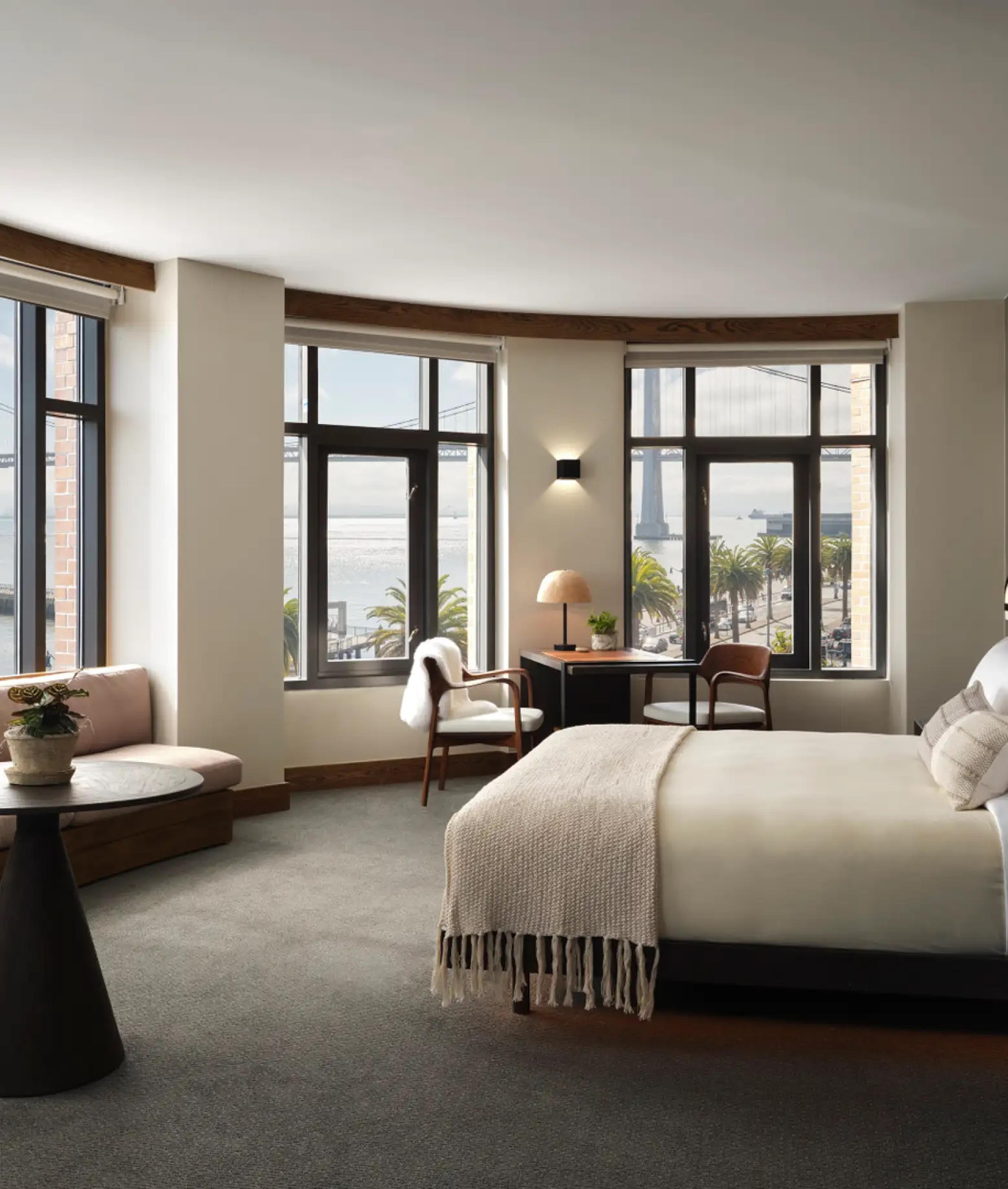 A corner room with a king bed in front of a curved window, a pale pink banquette, a round black table, and a desk, with the Bay Bridge and palm trees outside.