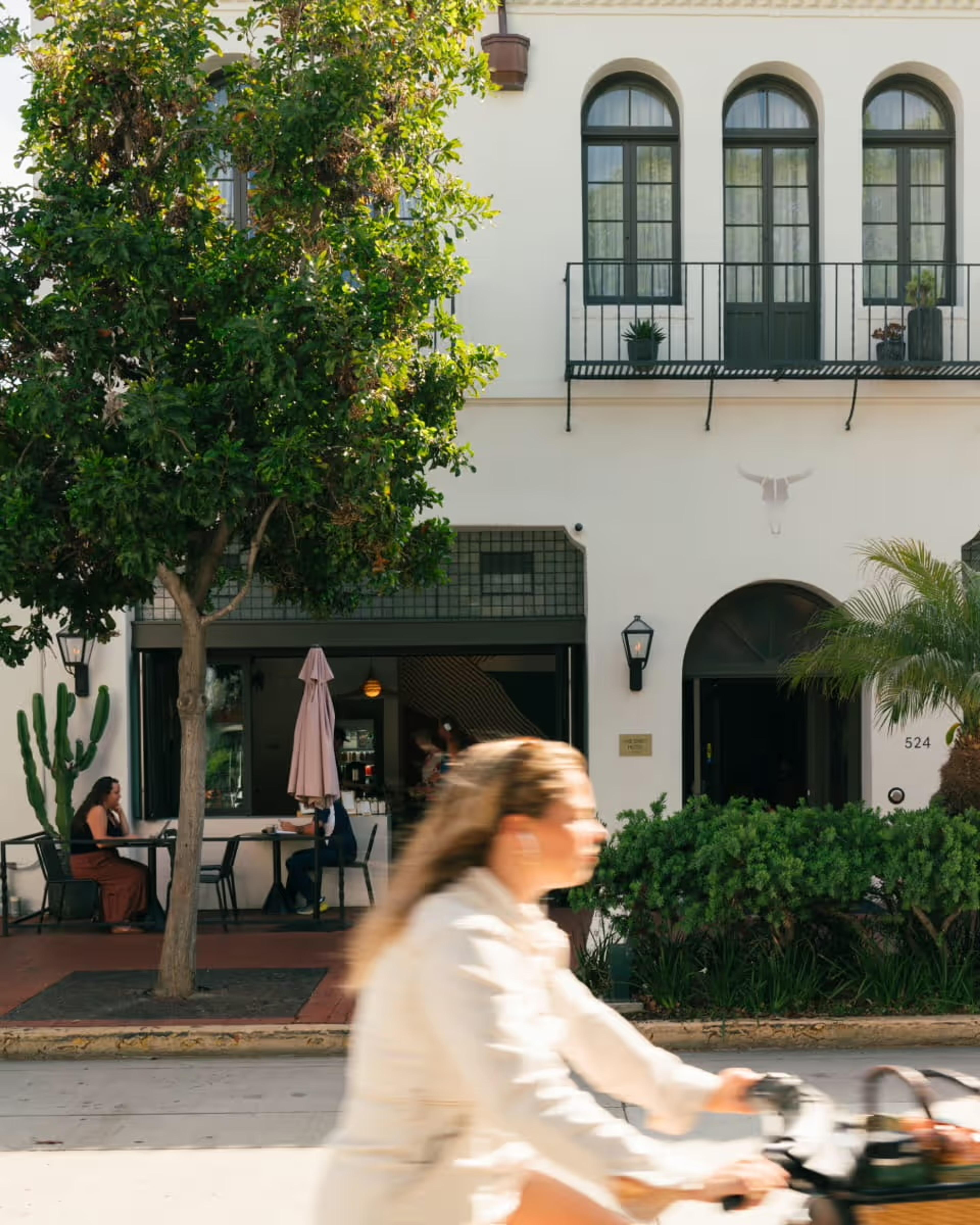 A woman cycles past a white Spanish colonial style hotel building with arched windows, wrought iron balconies, and a street level cafe with people seated under a tree.