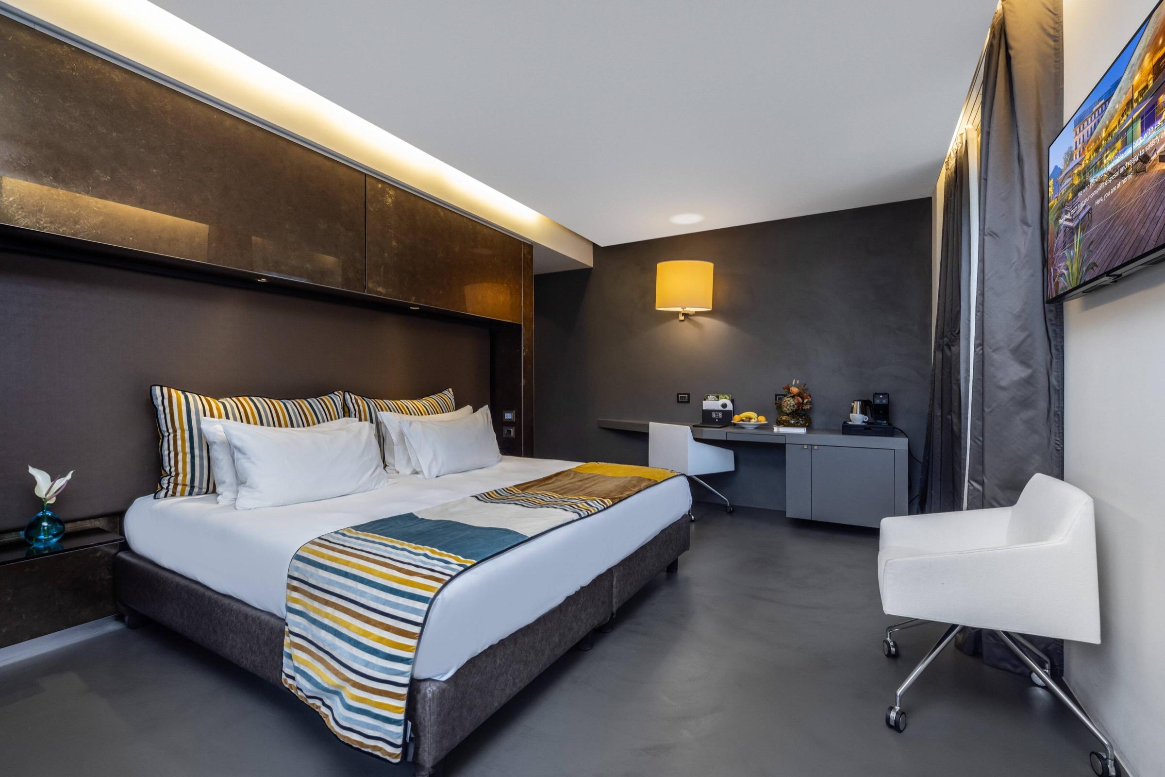 A hotel bedroom with a dark upholstered headboard, a bed dressed in a striped yellow and blue throw, and a white desk chair by the window.