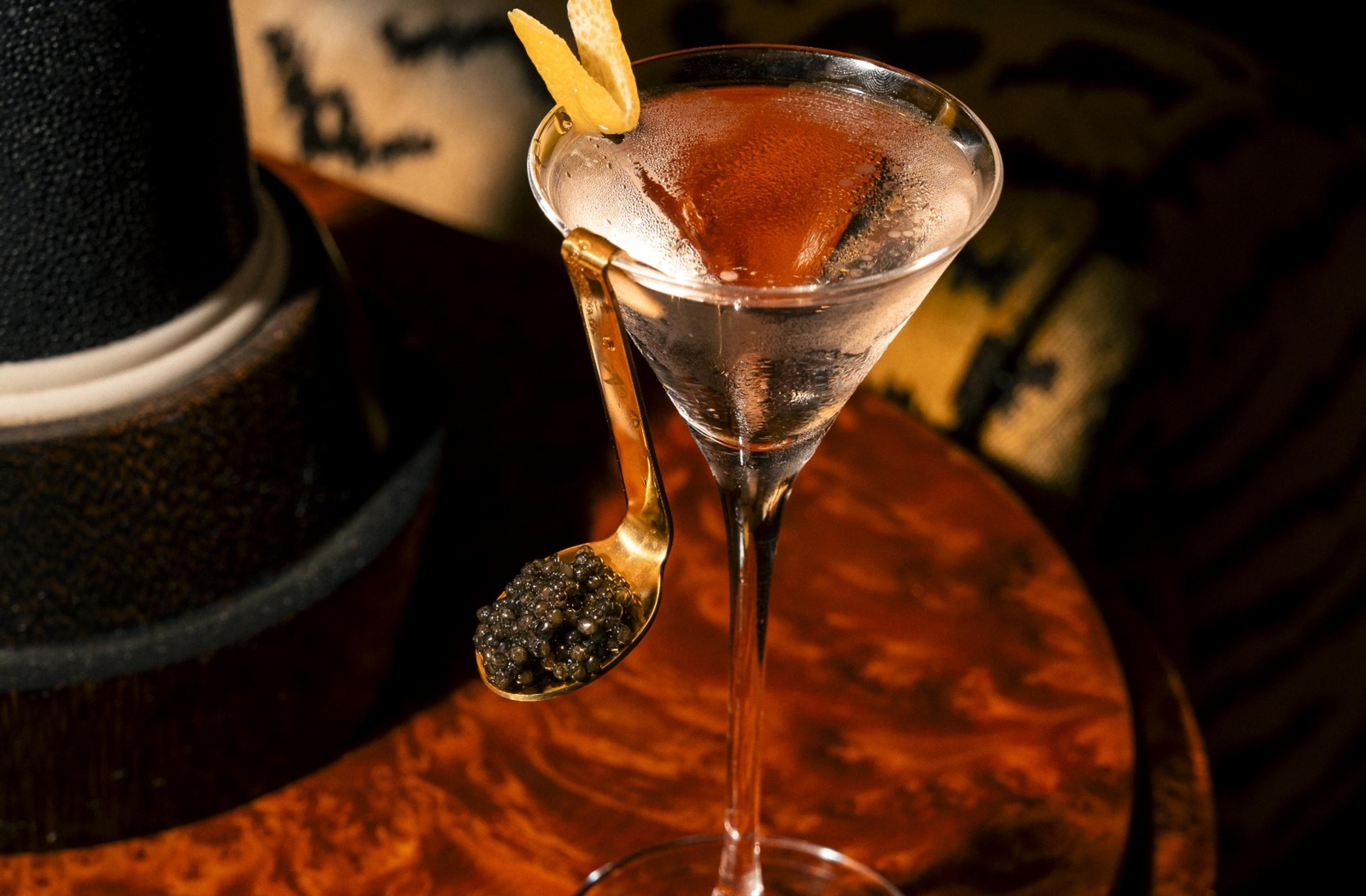 A martini glass with a lemon twist and a caviar-filled gold spoon hooked on its rim, with a black top hat behind on a wooden table.