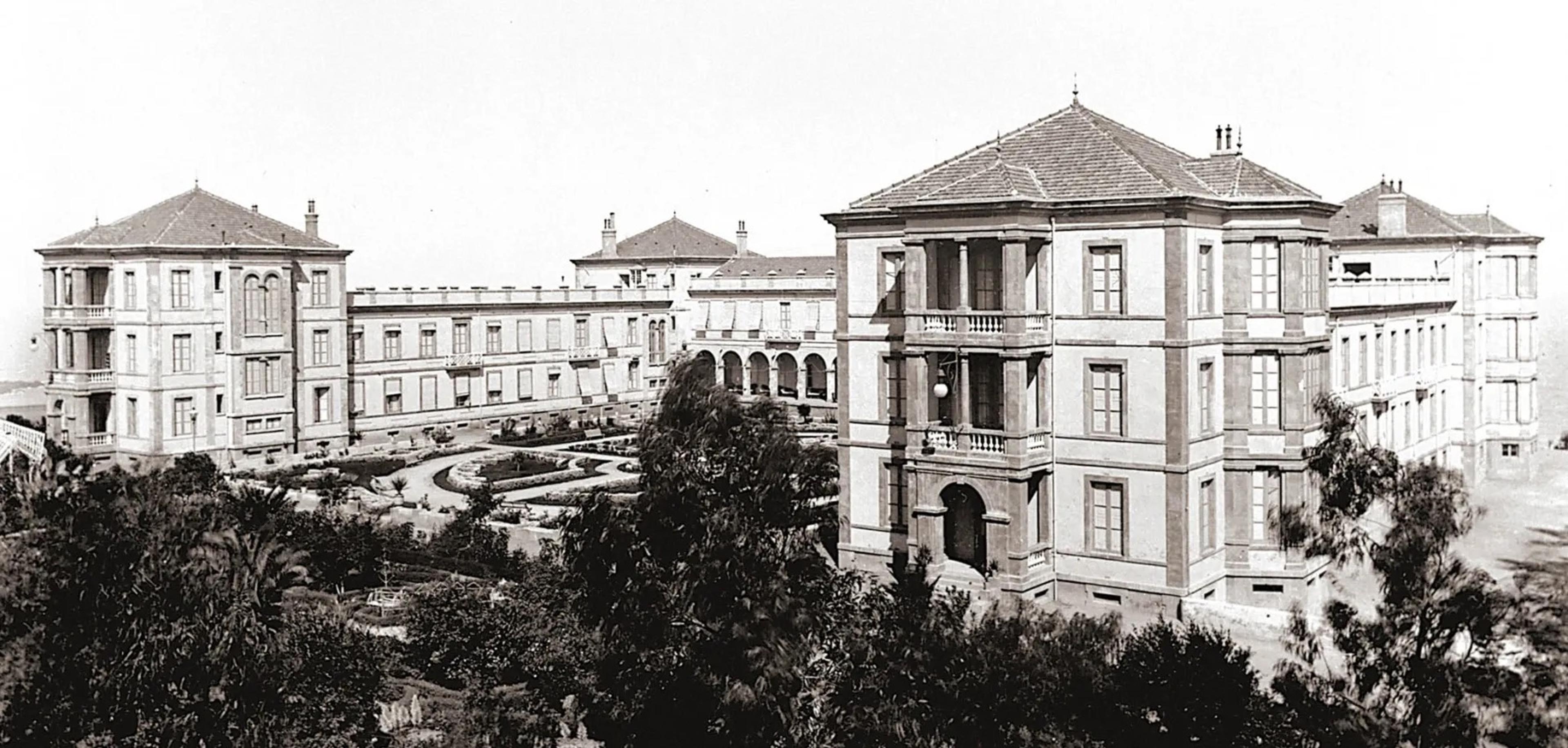 A black and white archival photograph of a grand hotel building with balconies and corner towers, seen past gardens and palm trees.