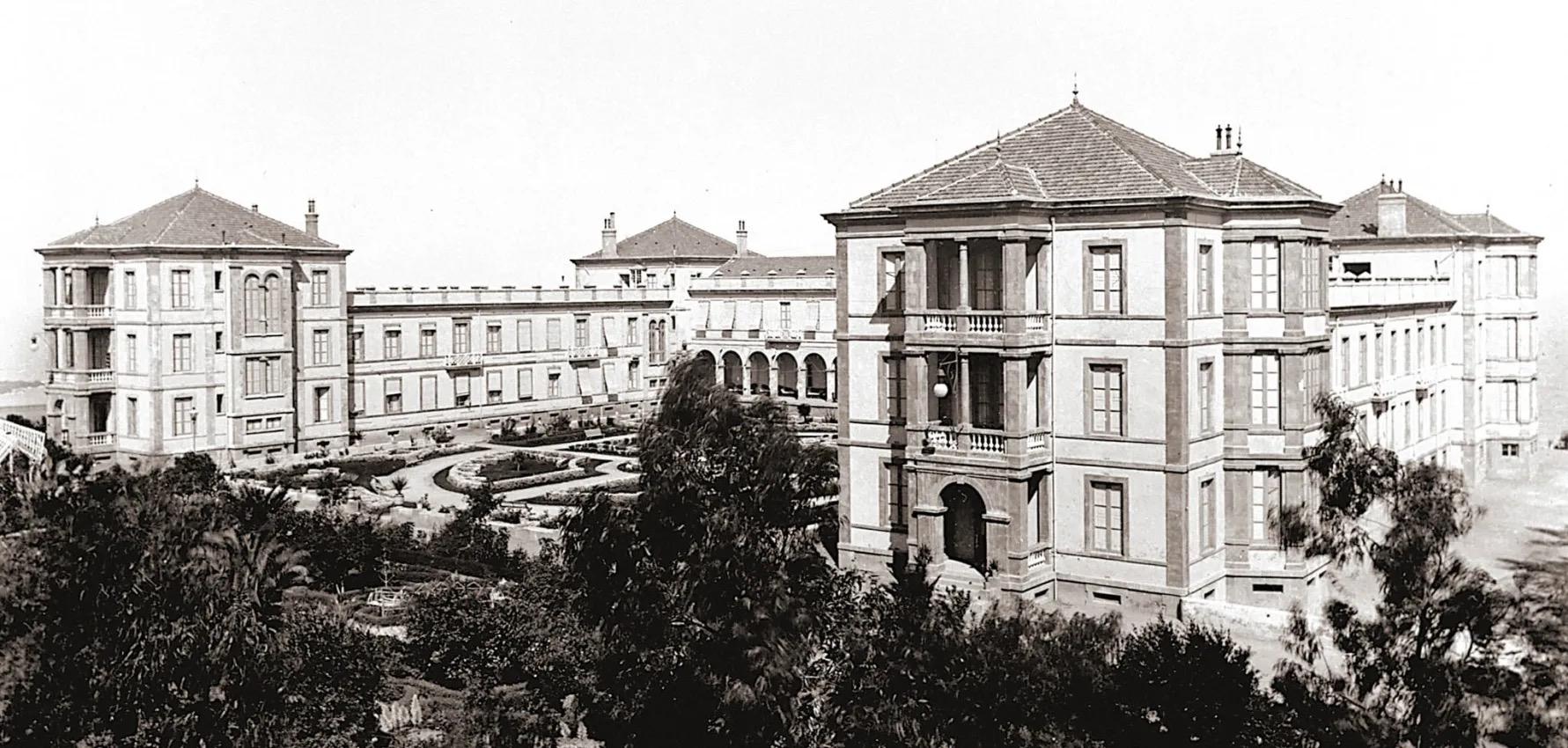 A black and white archival photograph of a grand hotel building with balconies and corner towers, seen past gardens and palm trees.