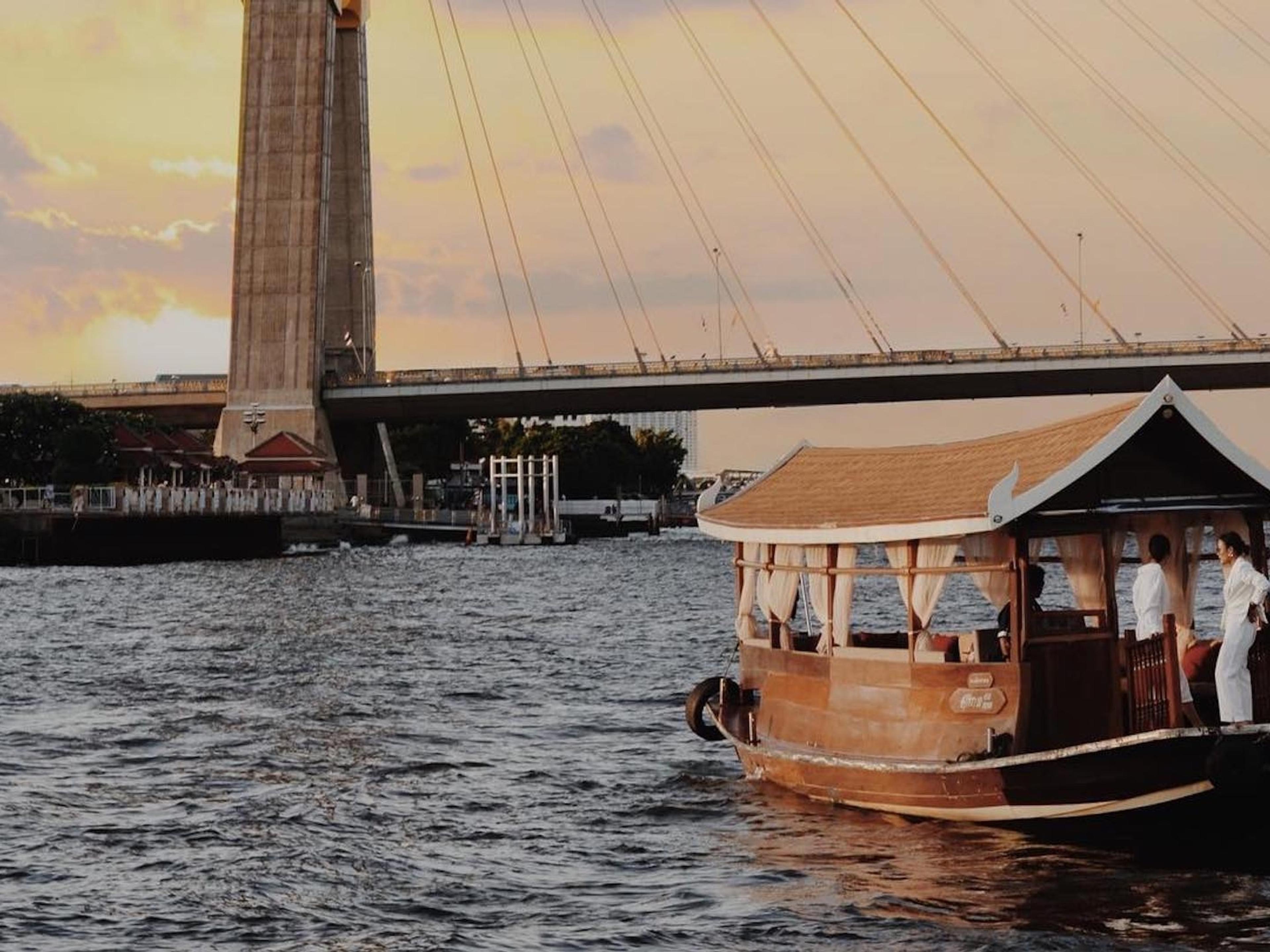 A wooden river boat with a peaked roof on the Chao Phraya at sunset, beneath a cable-stayed bridge