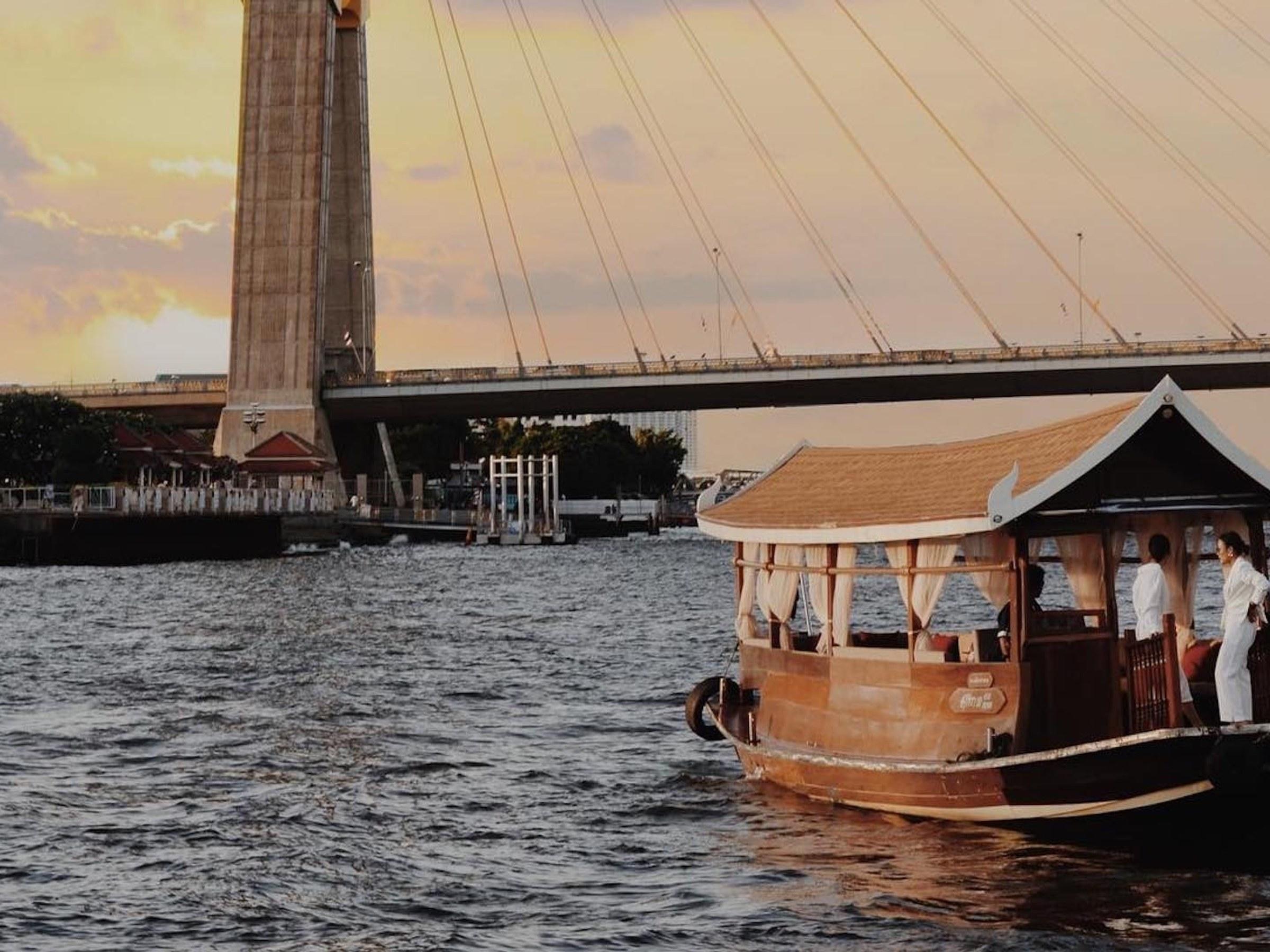 A wooden river boat with a peaked roof on the Chao Phraya at sunset, beneath a cable-stayed bridge