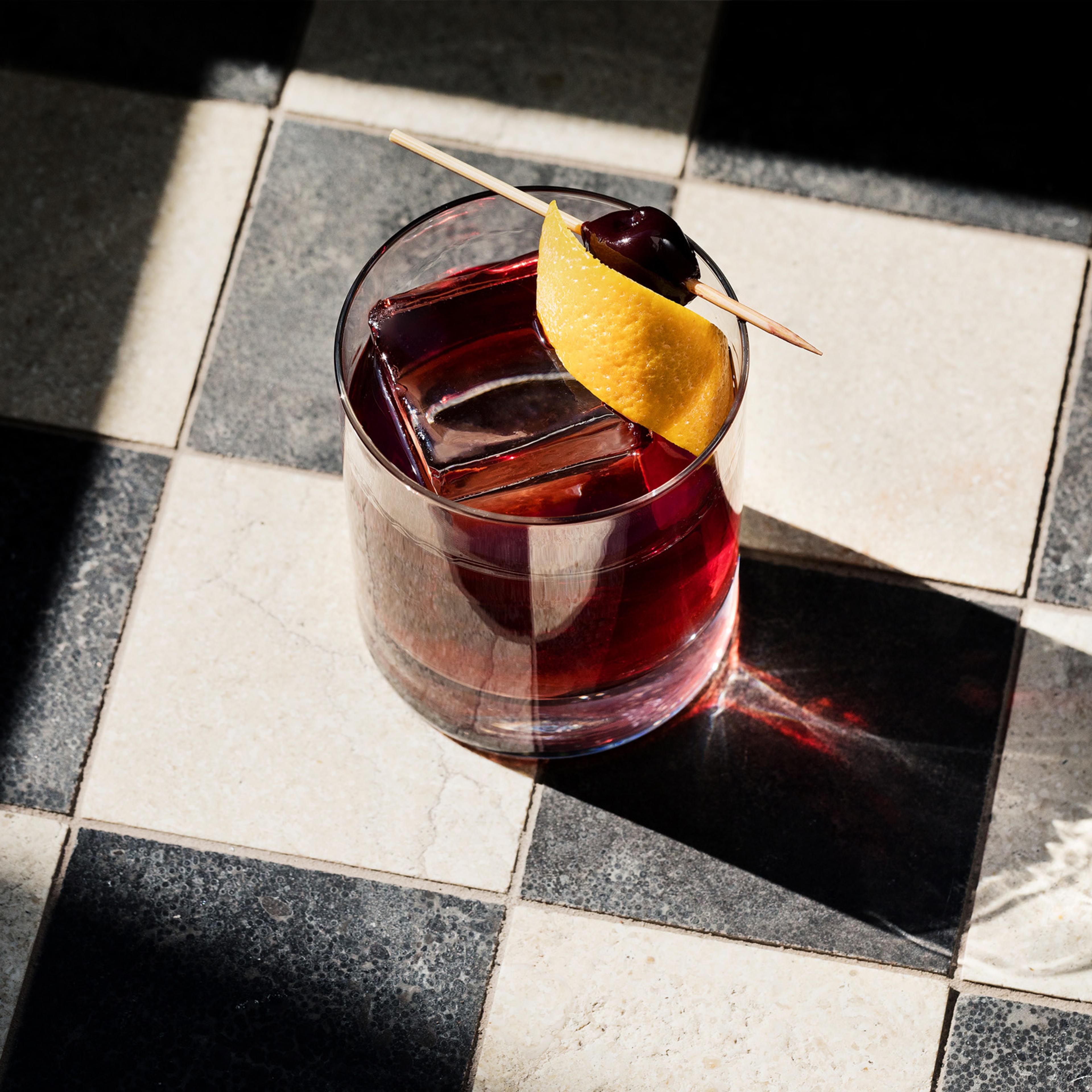 A dark cocktail garnished with an orange twist and a cherry on a pick, sitting in sunlight on a black and white tiled table.