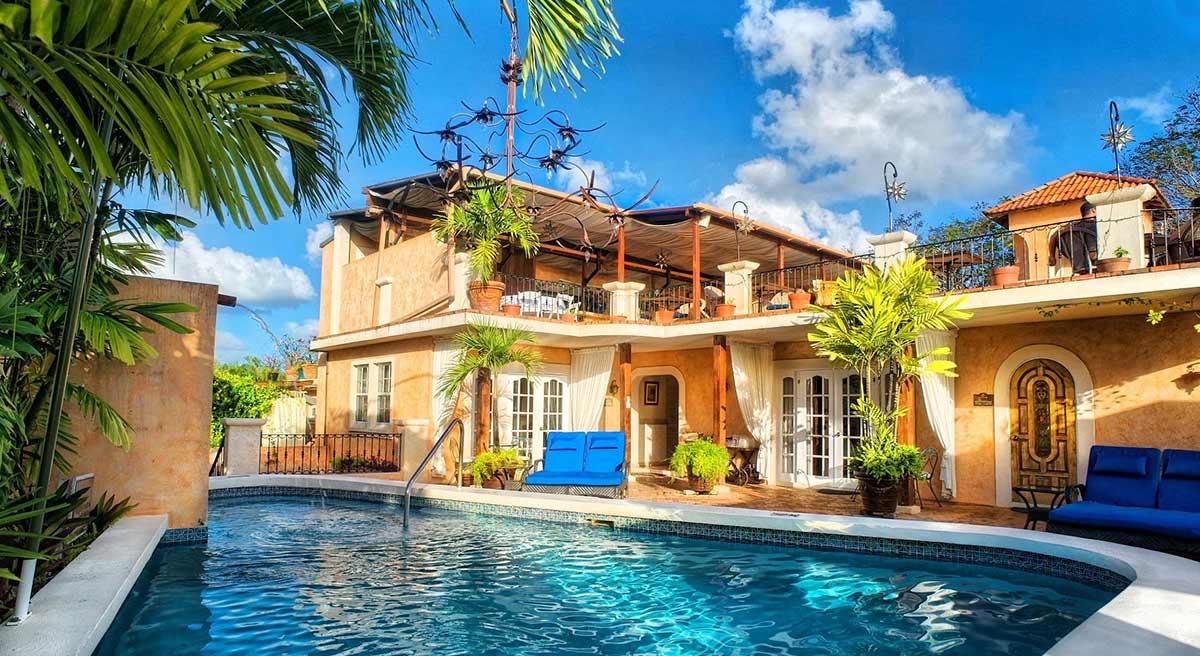 The hotel building seen from across its swimming pool, with balconies, French doors and palm trees against a blue sky.