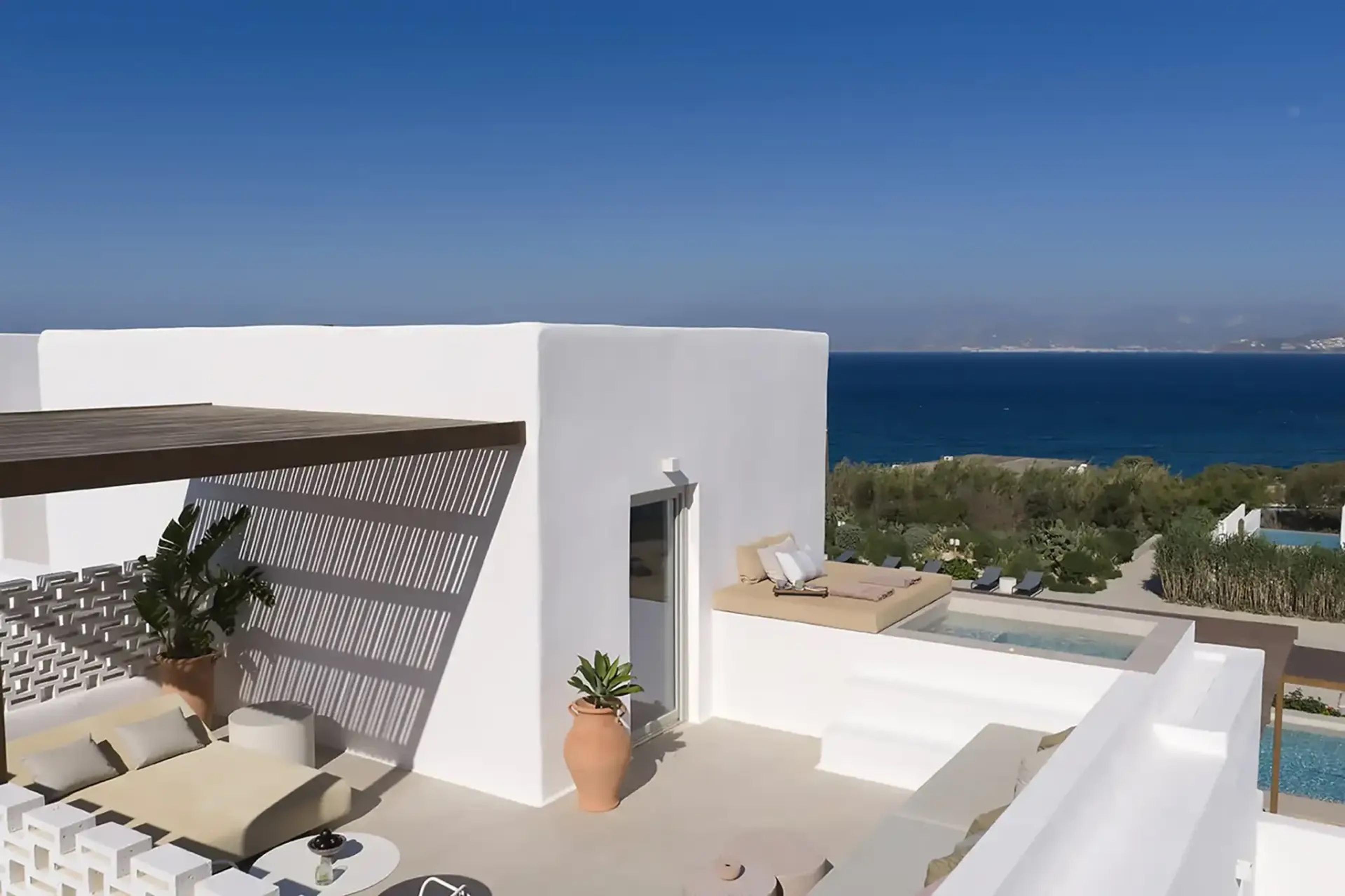 An aerial view of a whitewashed cube-shaped building with a small plunge pool and a sunbed on its terrace, the sea behind.