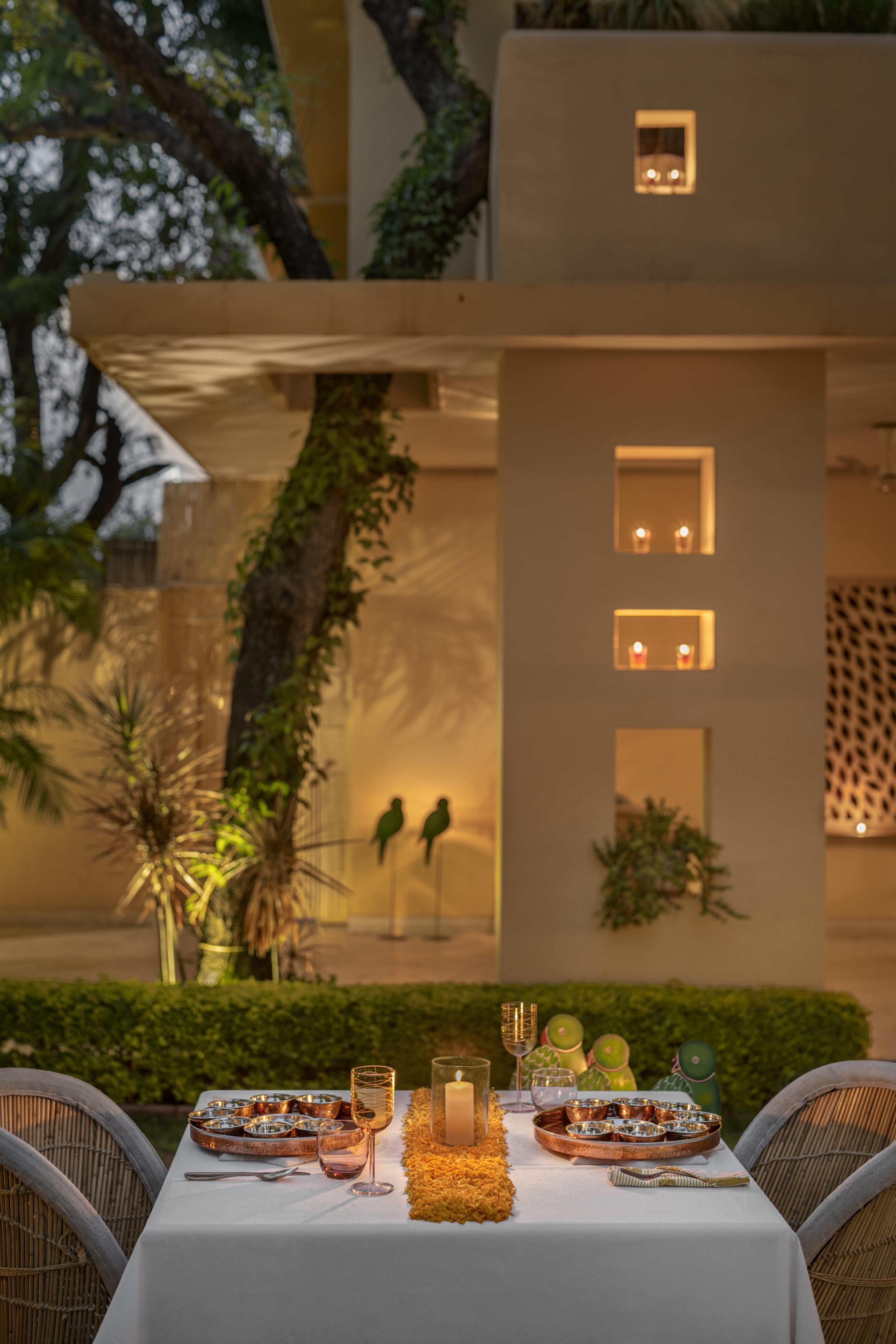 A table laid with a white cloth, wine glasses and a candle in front of a building lit up at dusk, with two parrot statues on the terrace.