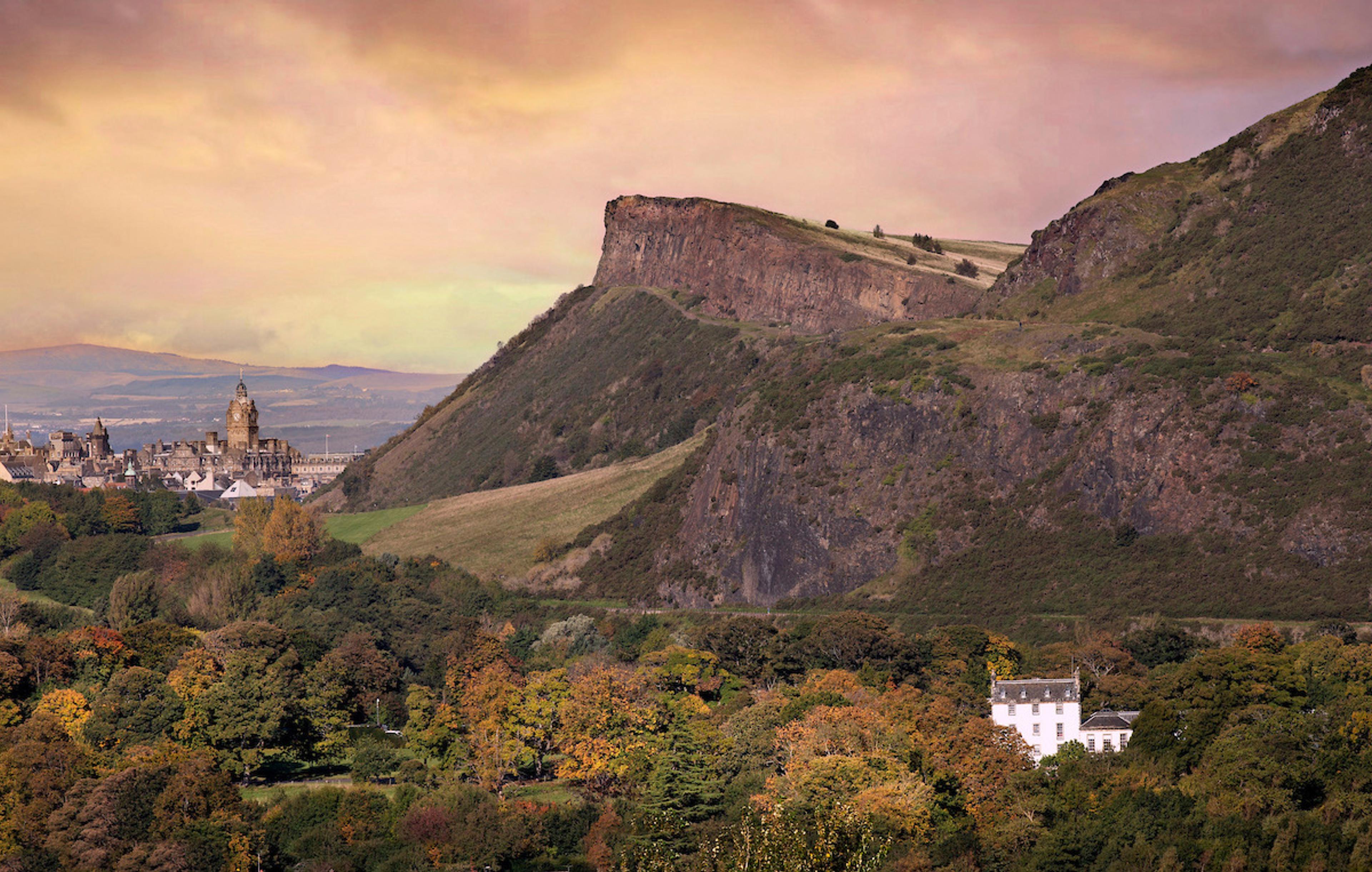 an aerial view of Prestonfield House's white facade among trees, with the cliffs of Salisbury Crags and Edinburgh's skyline behind it at dusk