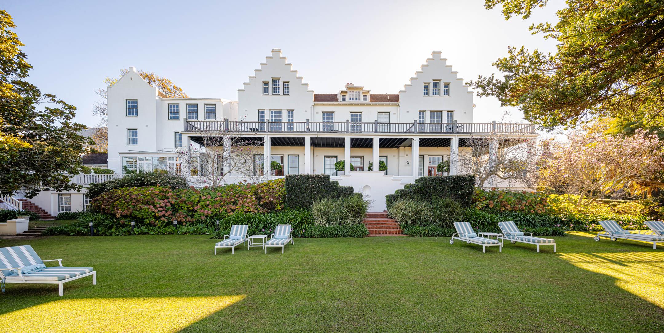 The back of a white building with a deep first-floor balcony, seen across a lawn with striped loungers.