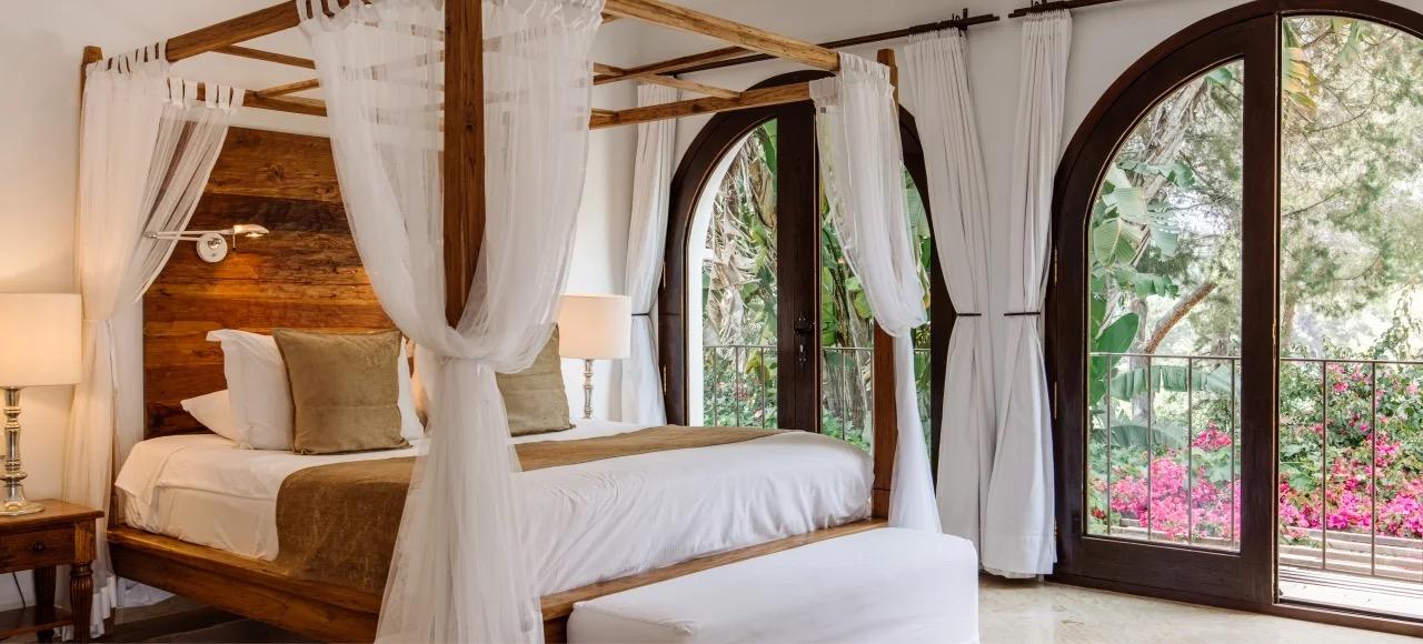 A four poster bed against a dark wood headboard, with tall arched doors open onto a garden of pink bougainvillea.