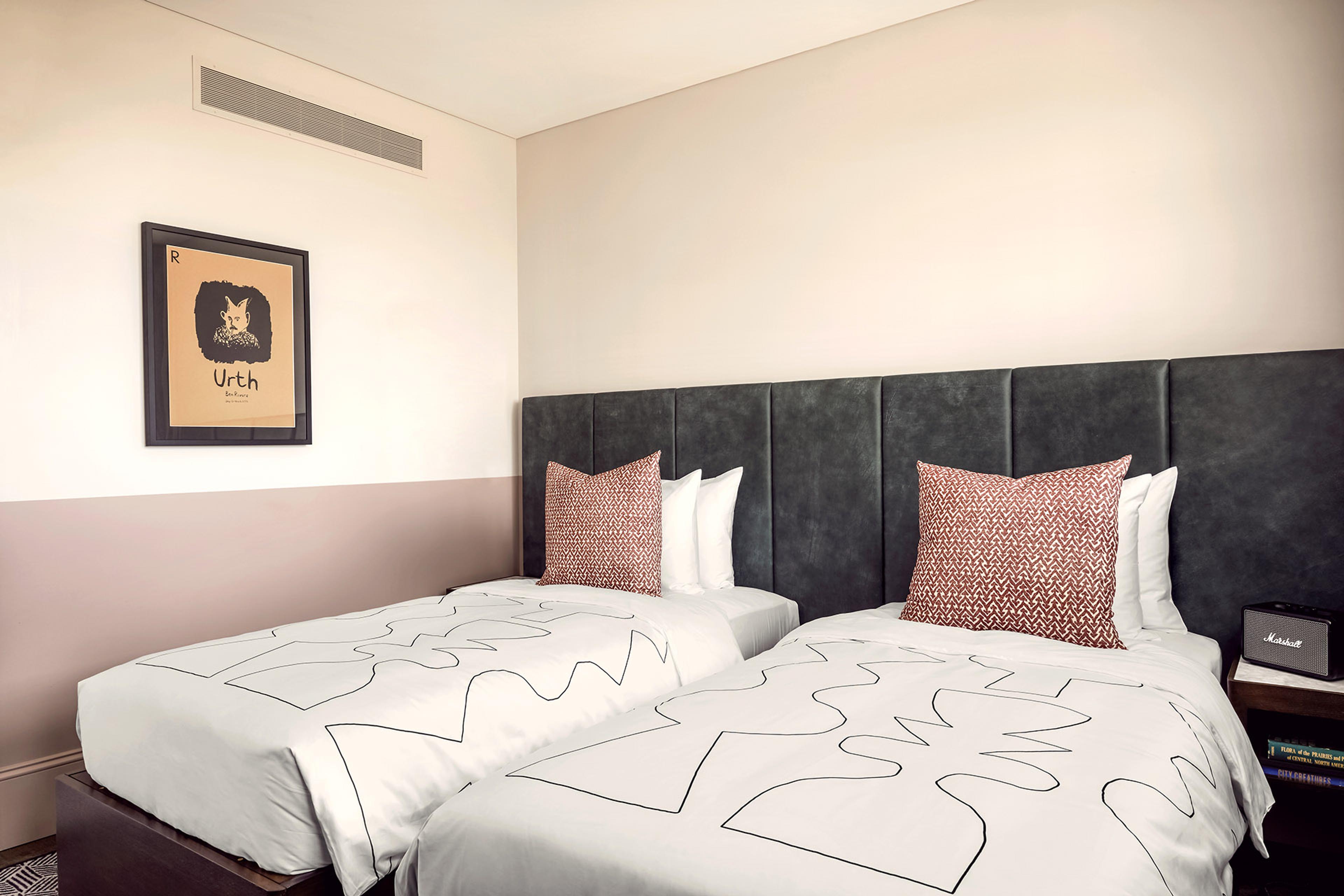 Two single beds side by side with a dark grey headboard, each with a patterned red and white cushion, under a framed print reading Urth.
