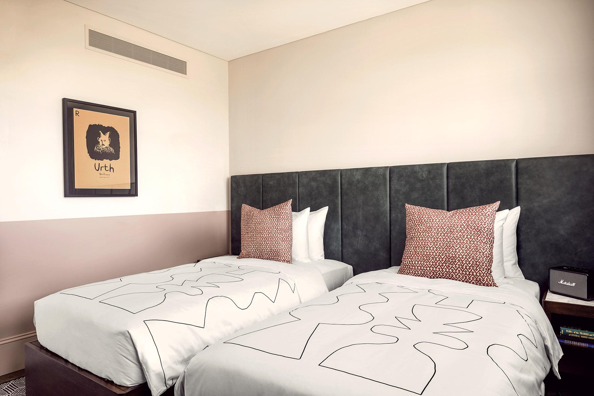 Two single beds side by side with a dark grey headboard, each with a patterned red and white cushion, under a framed print reading Urth.