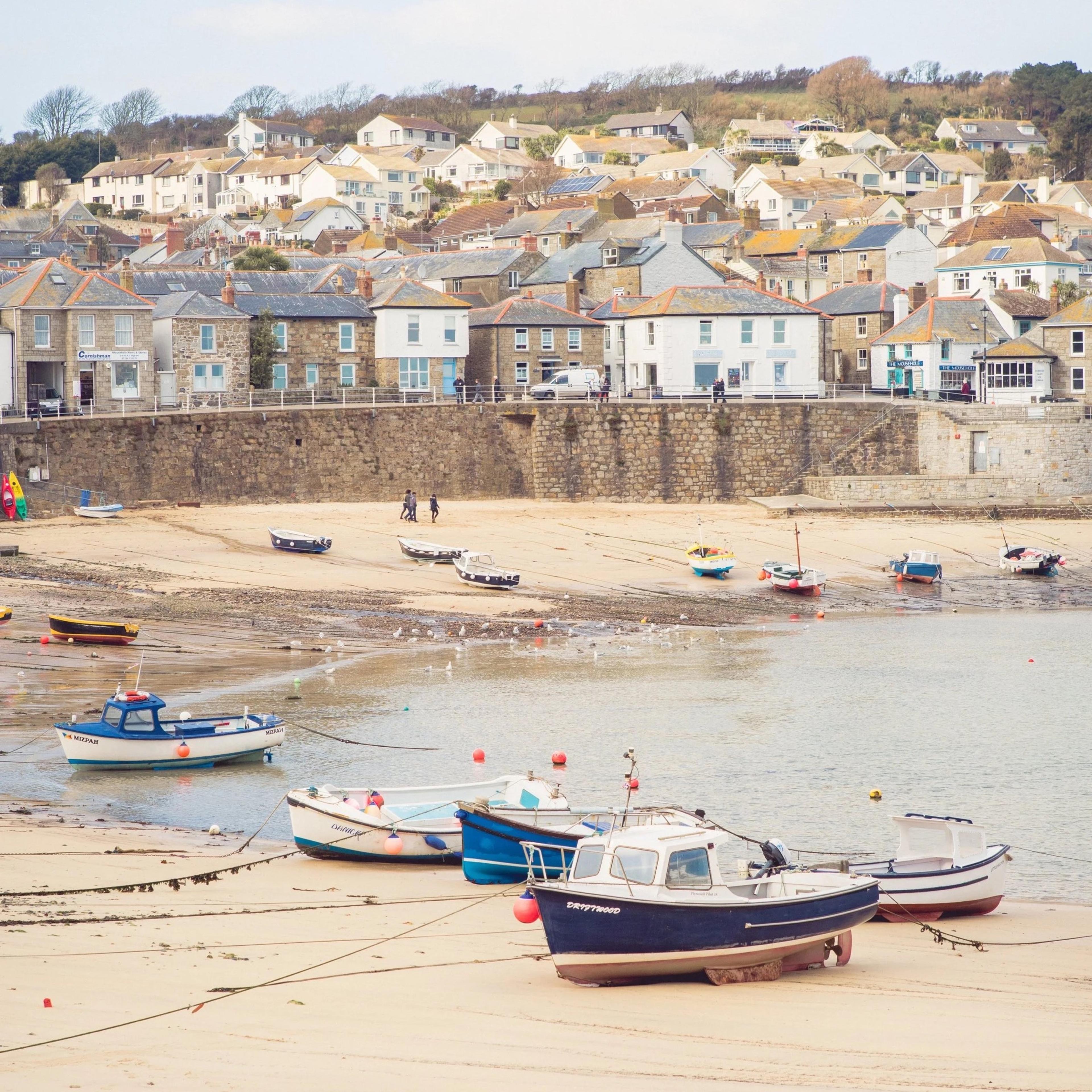 Newlyn harbour at low tide, with small fishing boats resting on the sand in front of a stone harbour wall and a hillside of houses.