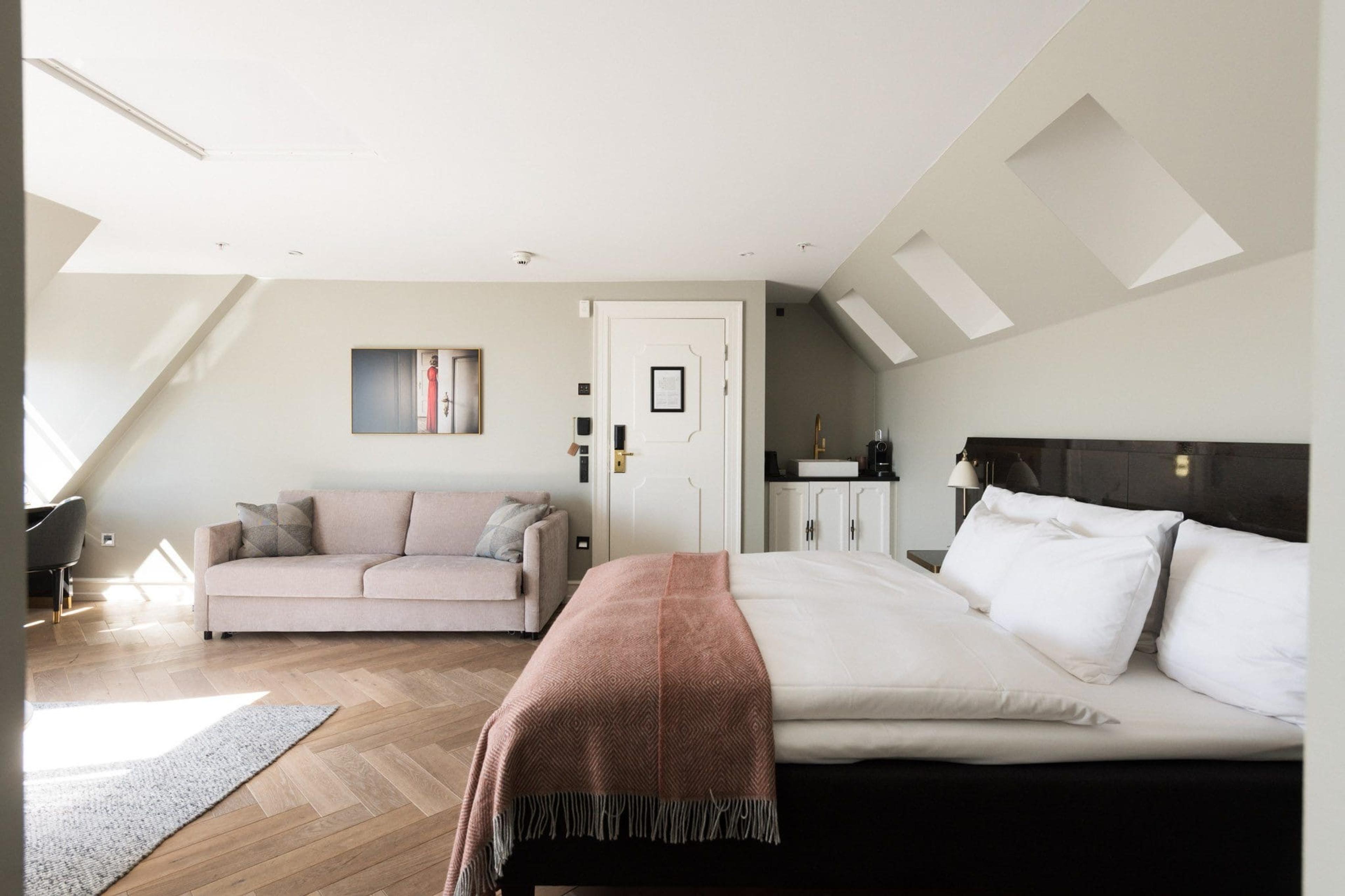 A loft bedroom with a dark wooden bed topped with a rose-coloured throw, a pale pink sofa, and a sloped ceiling with skylights over a herringbone wood floor.