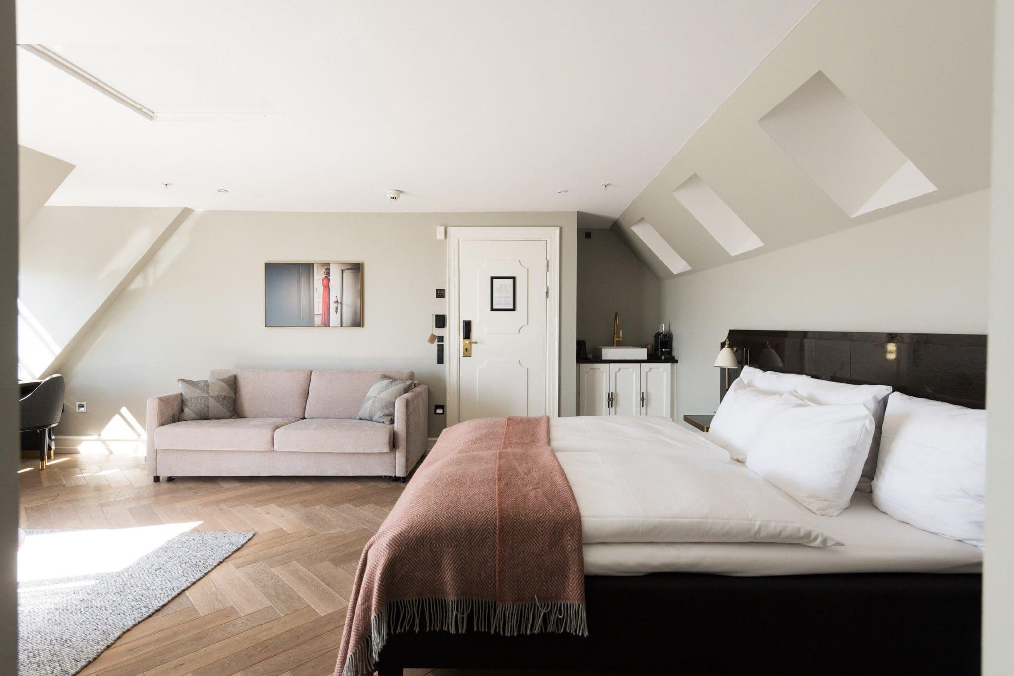 A loft bedroom with a dark wooden bed topped with a rose-coloured throw, a pale pink sofa, and a sloped ceiling with skylights over a herringbone wood floor.