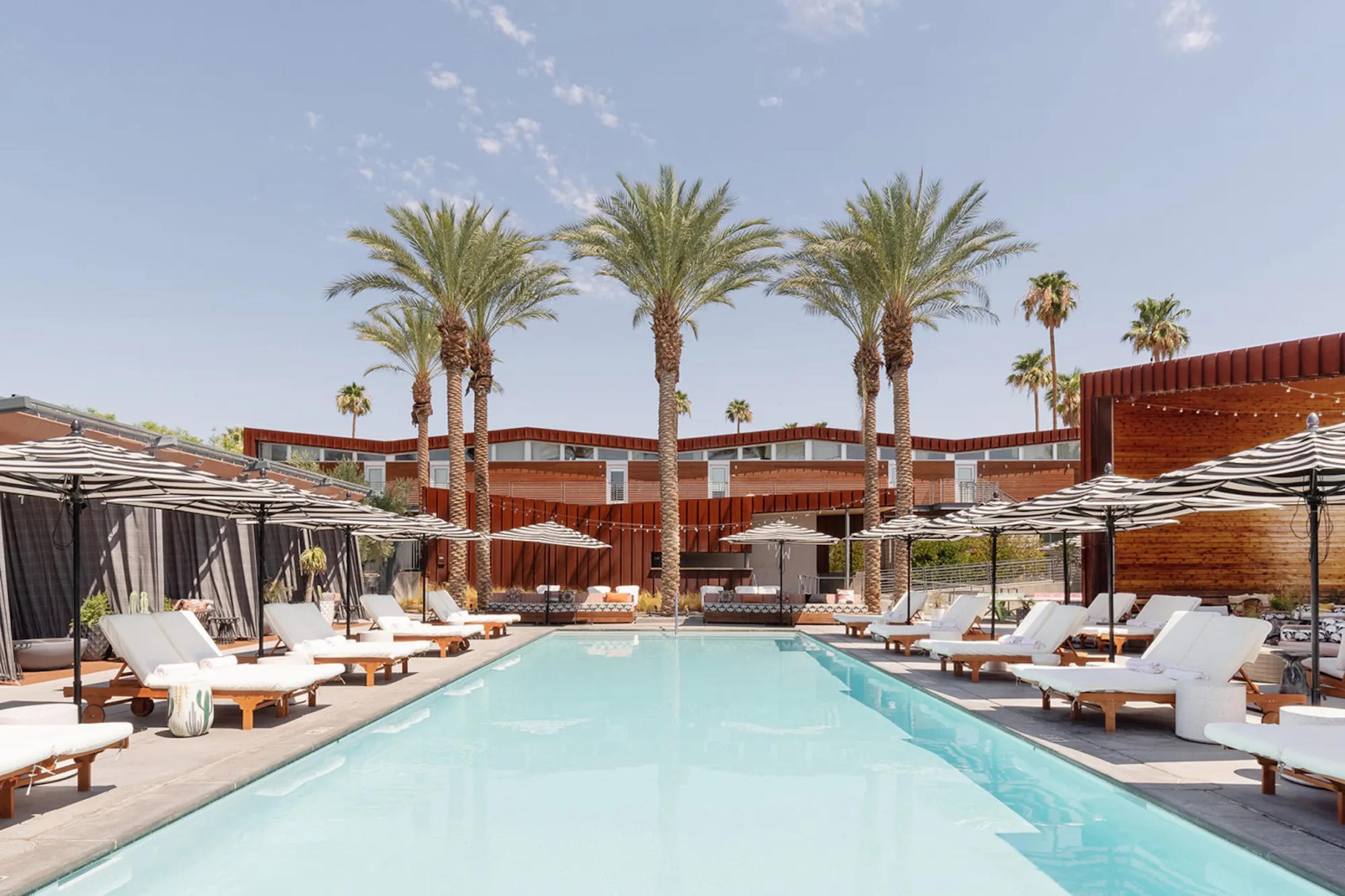 A long rectangular swimming pool with pale blue water, edged by white-cushioned sun loungers under striped parasols and tall palm trees, with the rust-coloured building behind.