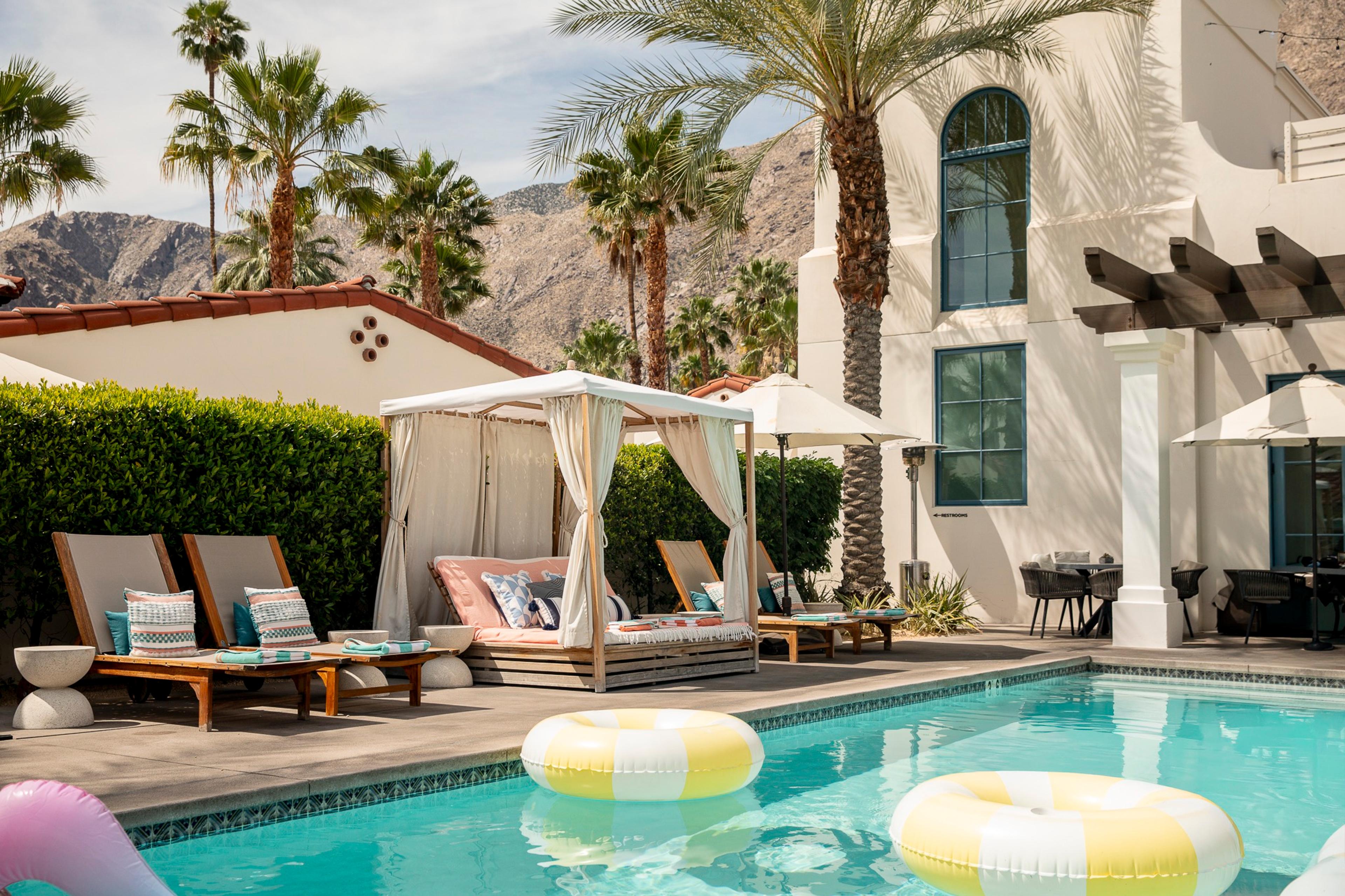 A rectangular swimming pool with two striped inflatable rings, wooden loungers and a white canopied daybed beside a building with arched blue-framed windows.