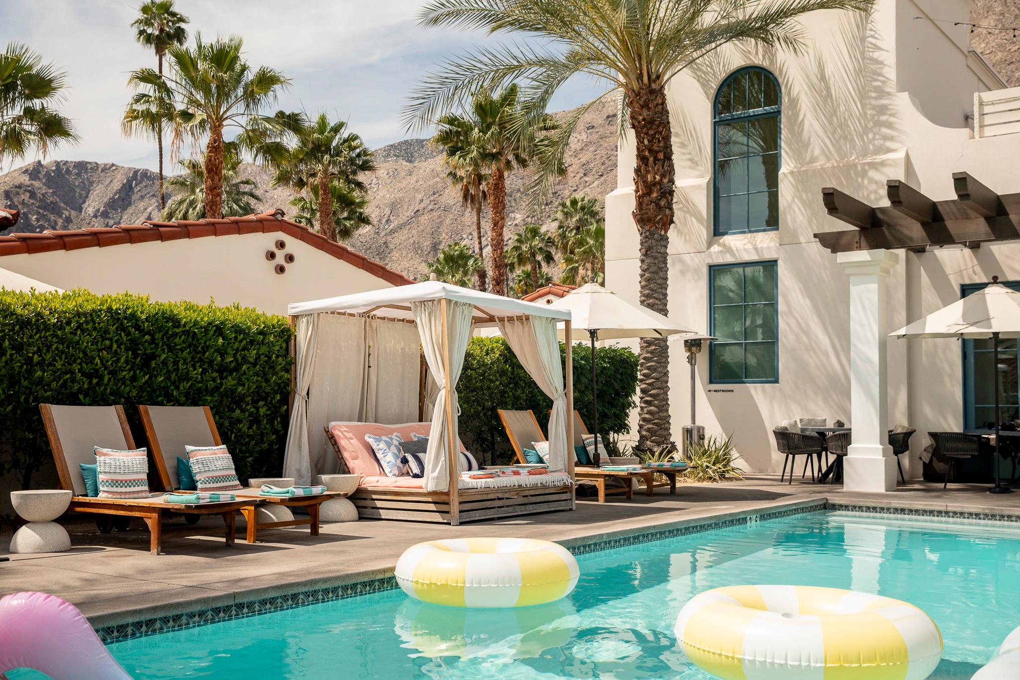 A rectangular swimming pool with two striped inflatable rings, wooden loungers and a white canopied daybed beside a building with arched blue-framed windows.