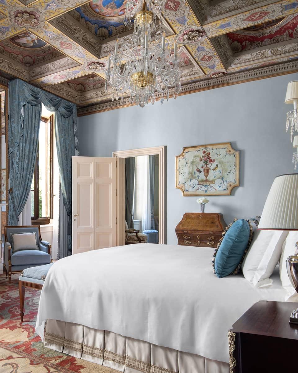 A bedroom with a painted coffered ceiling, a crystal chandelier, and a bed with white linens and a teal cushion against pale blue walls.