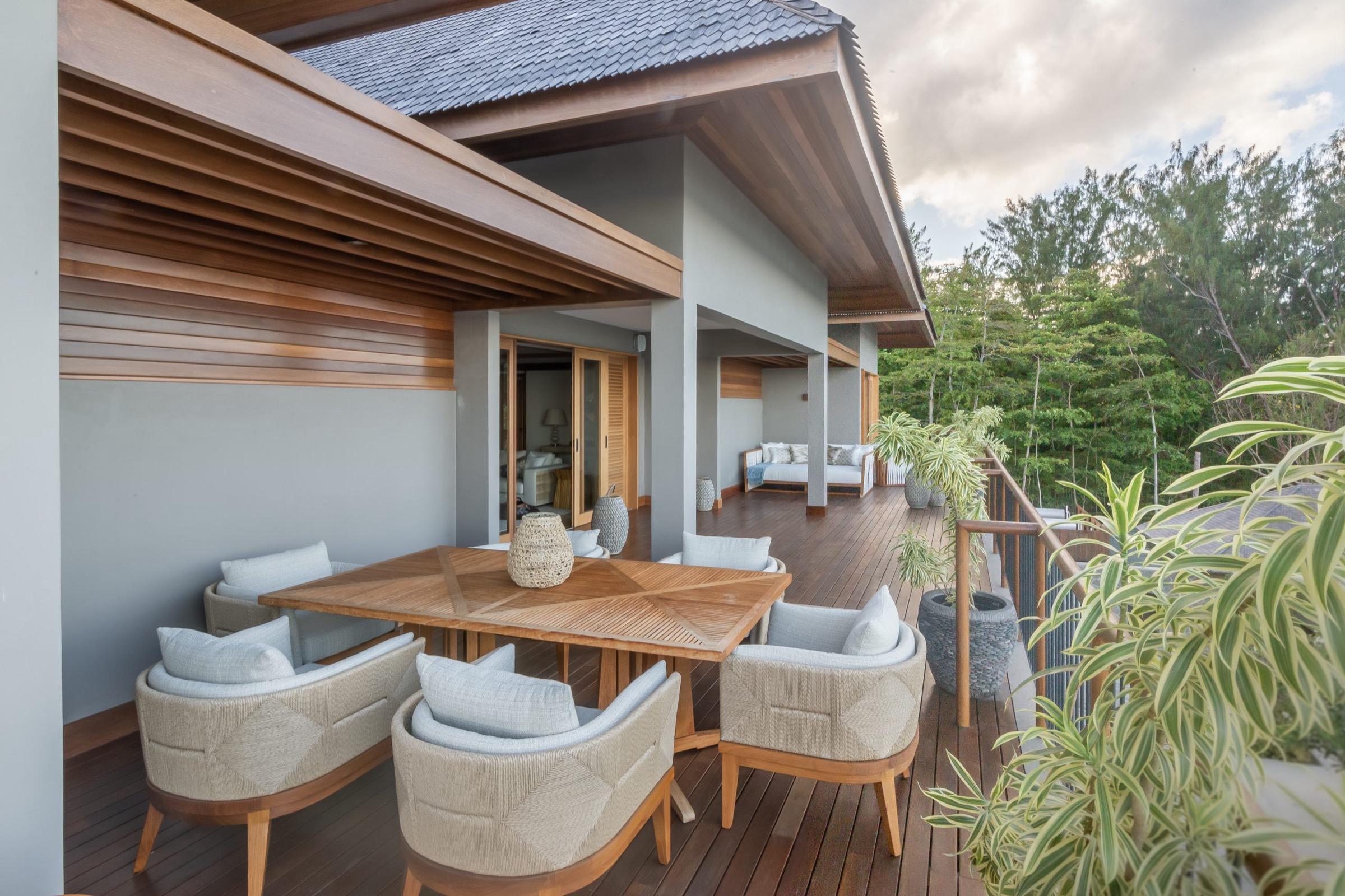 An outdoor dining table with woven chairs on a covered wooden deck, with a sofa beyond and forested hills in the distance.