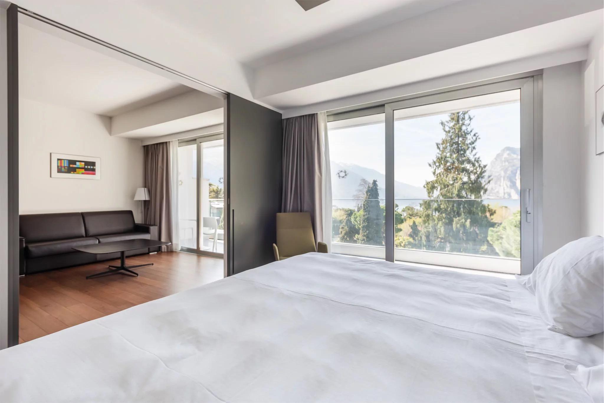 A bedroom with a made white bed in the foreground, a sliding door to a sitting room with a dark leather sofa, and tall windows onto trees and a rocky mountain above the lake.
