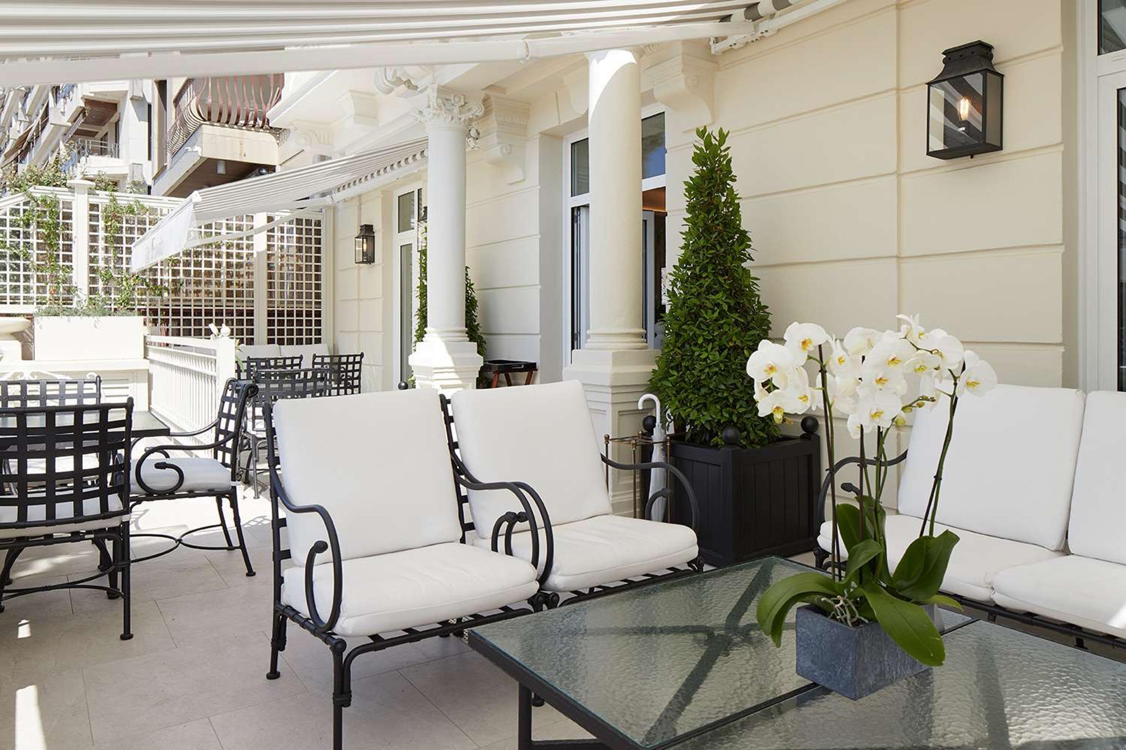 A covered rooftop terrace with wrought-iron armchairs and sofas in white cushions, topiary and orchids, and the hotel's own cream facade behind.