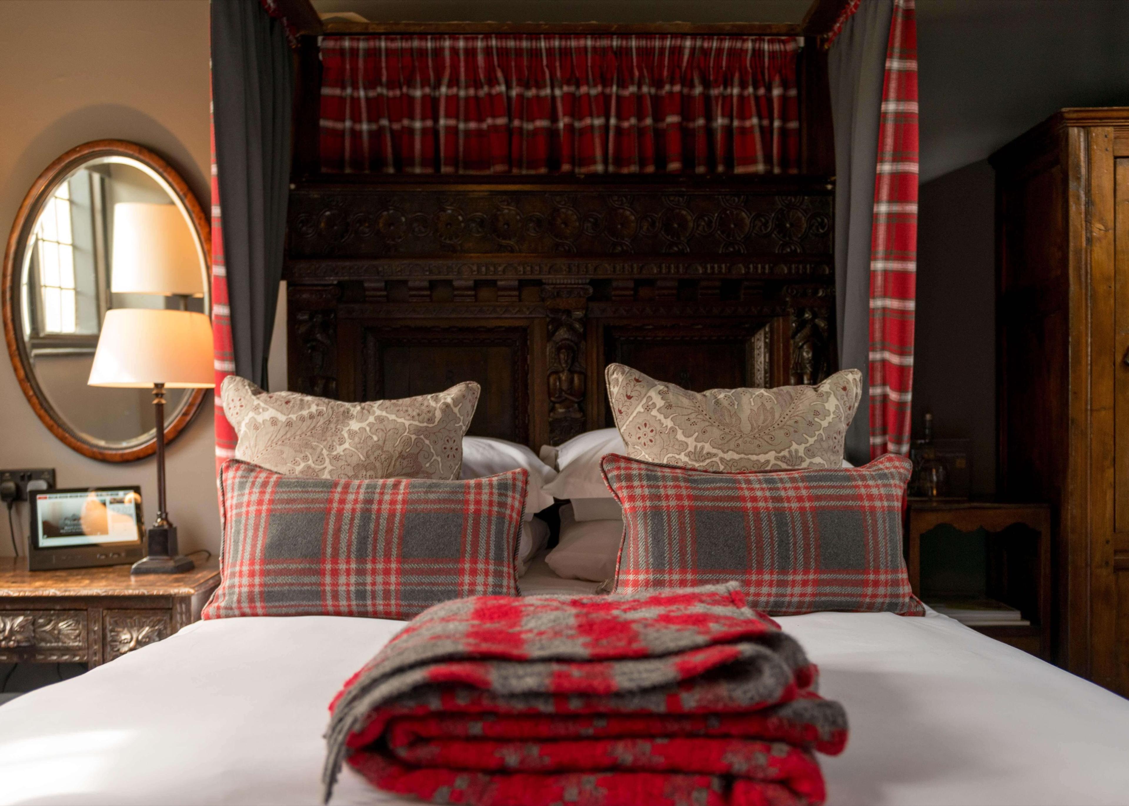A four-poster bed with dark carved wood and a scalloped tartan canopy, piled with tartan and paisley cushions and a folded tartan blanket, beside a round mirror and a lit lamp.