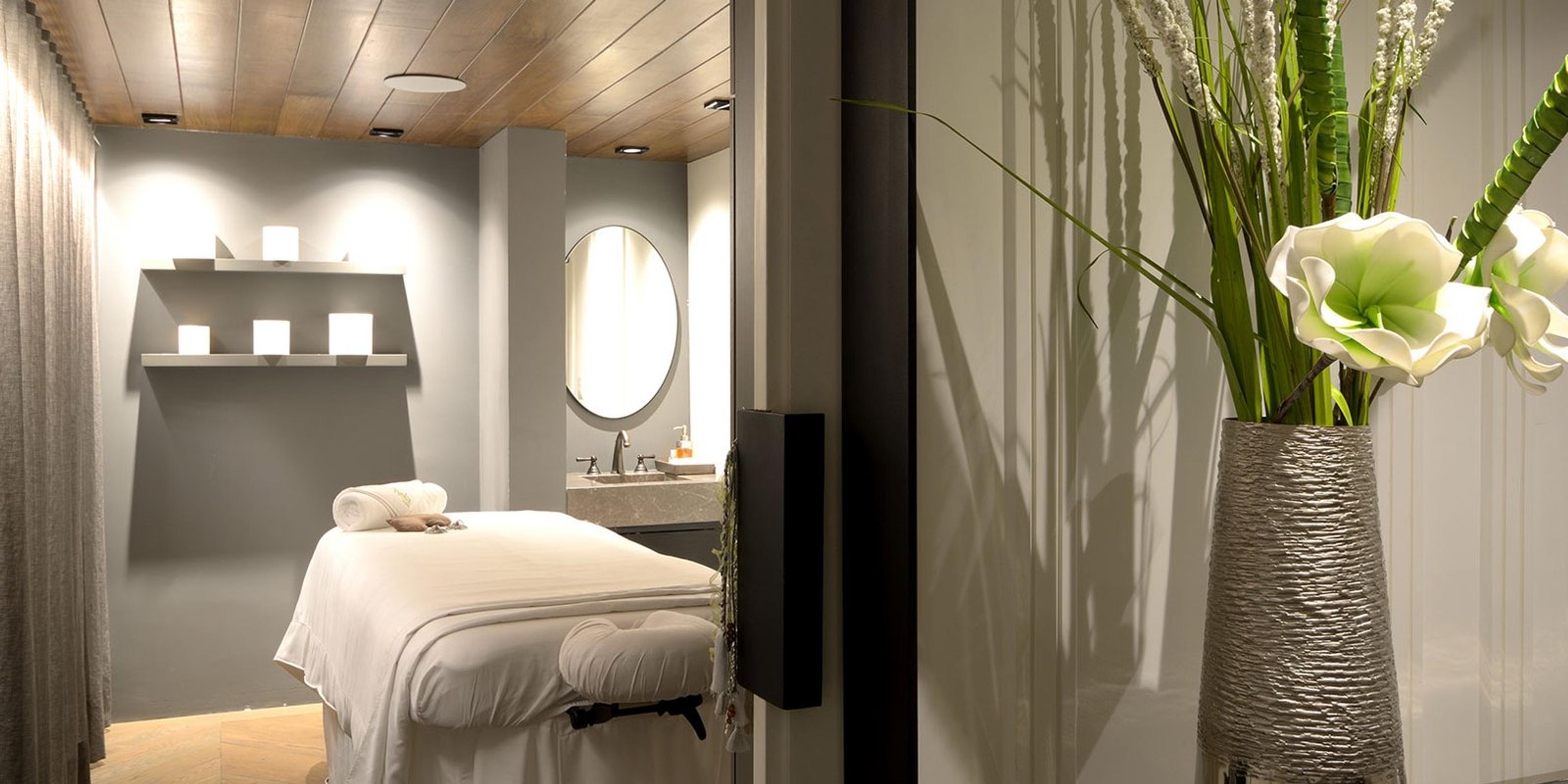An empty spa treatment room with a made-up massage table, a round mirror, candles on a shelf and a vase of white flowers in the foreground.