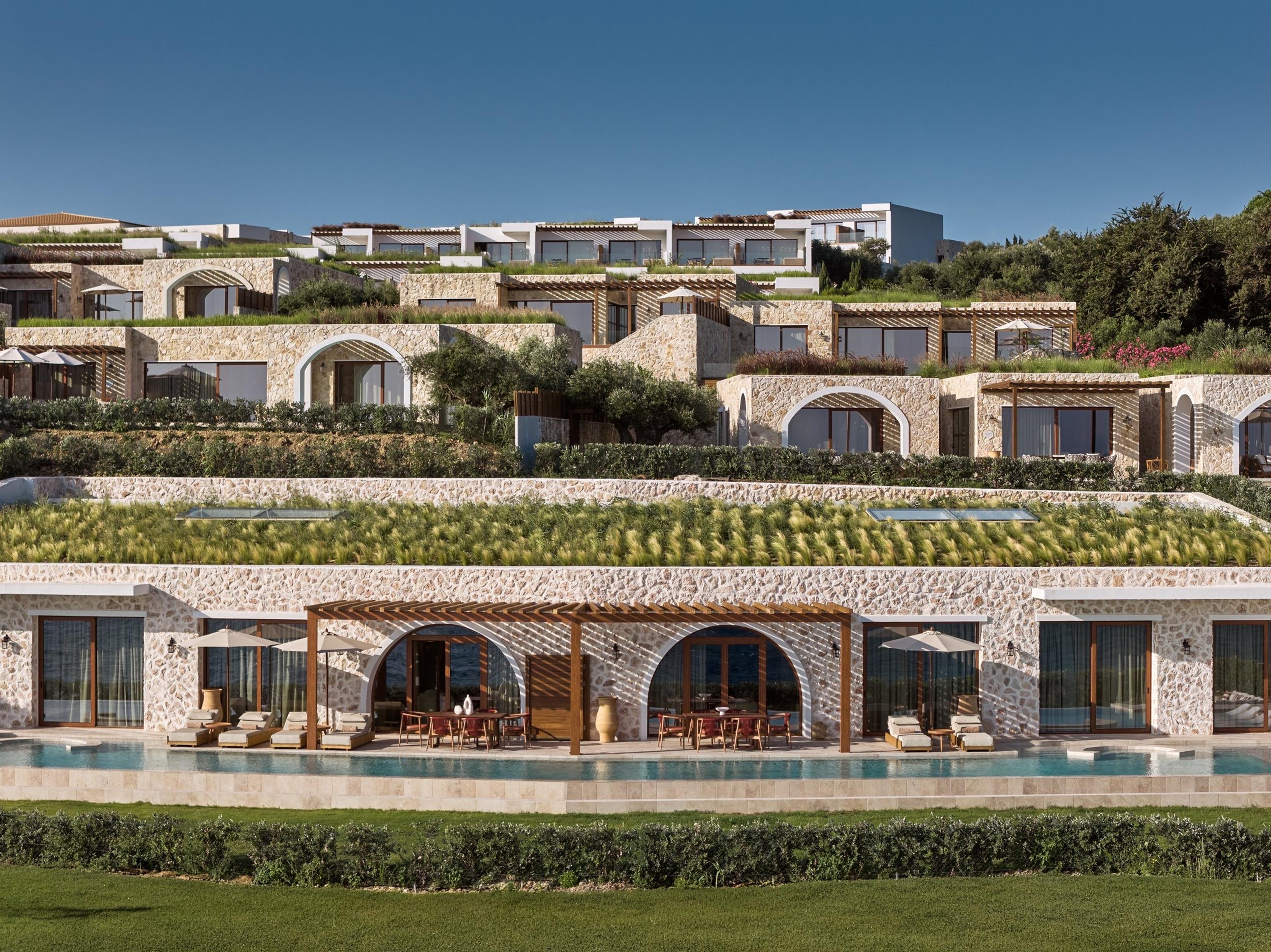 Aerial daylight view of a tiered stone and white building with a long pool and terrace in front, set among olive and cypress trees under a clear sky.