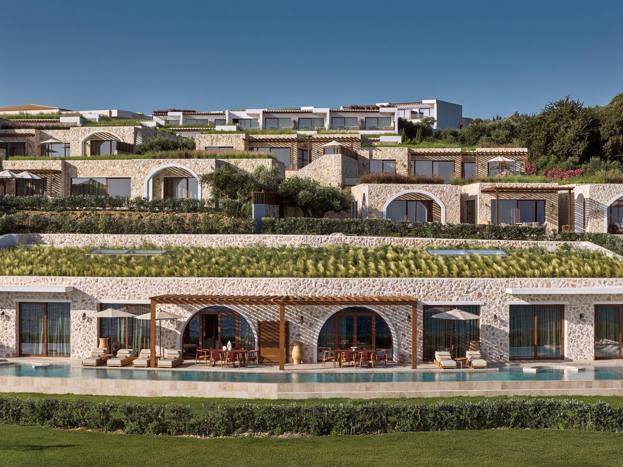 Aerial daylight view of a tiered stone and white building with a long pool and terrace in front, set among olive and cypress trees under a clear sky.