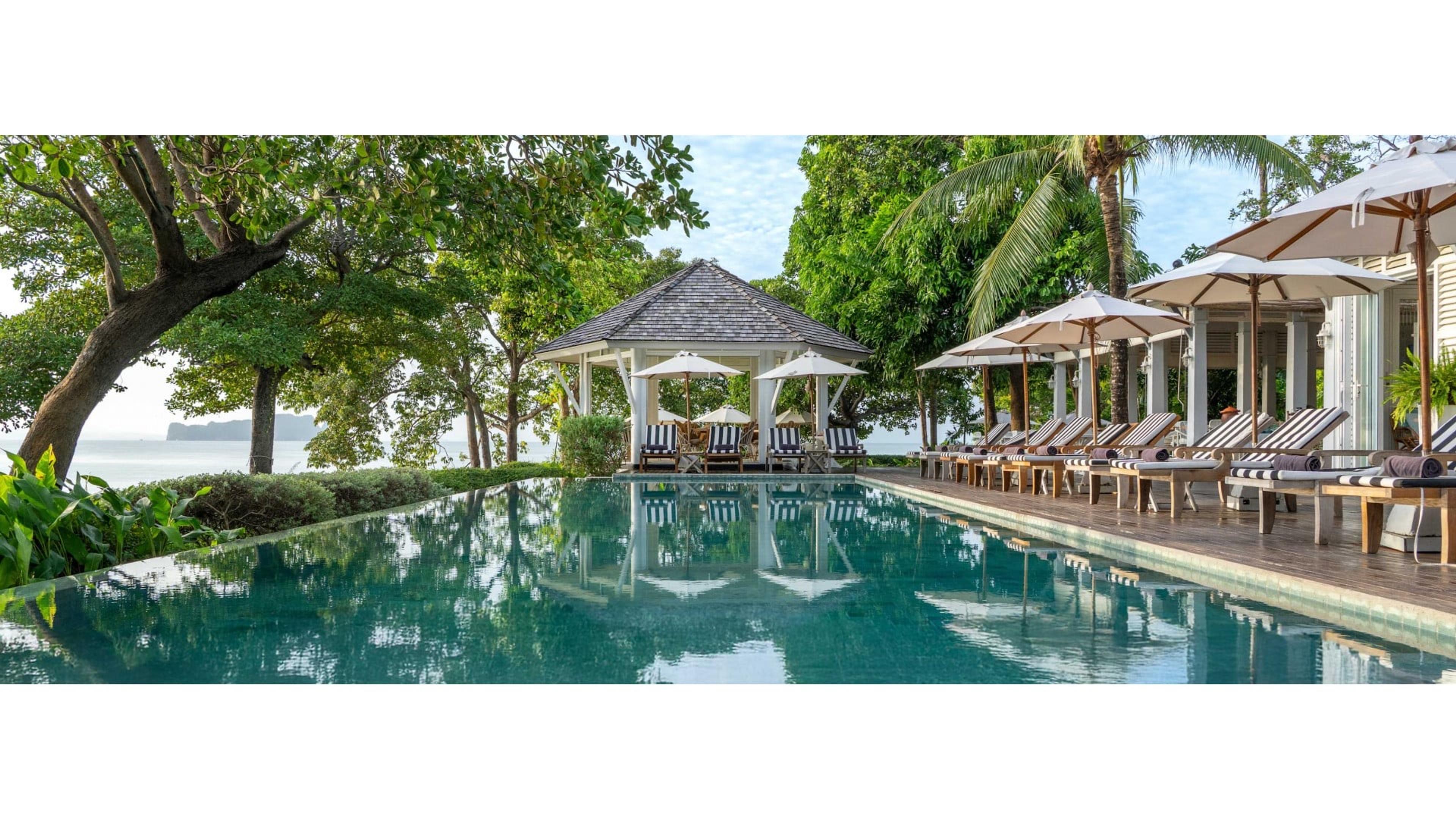 An infinity pool beside a thatched-roof gazebo and striped loungers under trees, looking out over the sea to limestone islands.