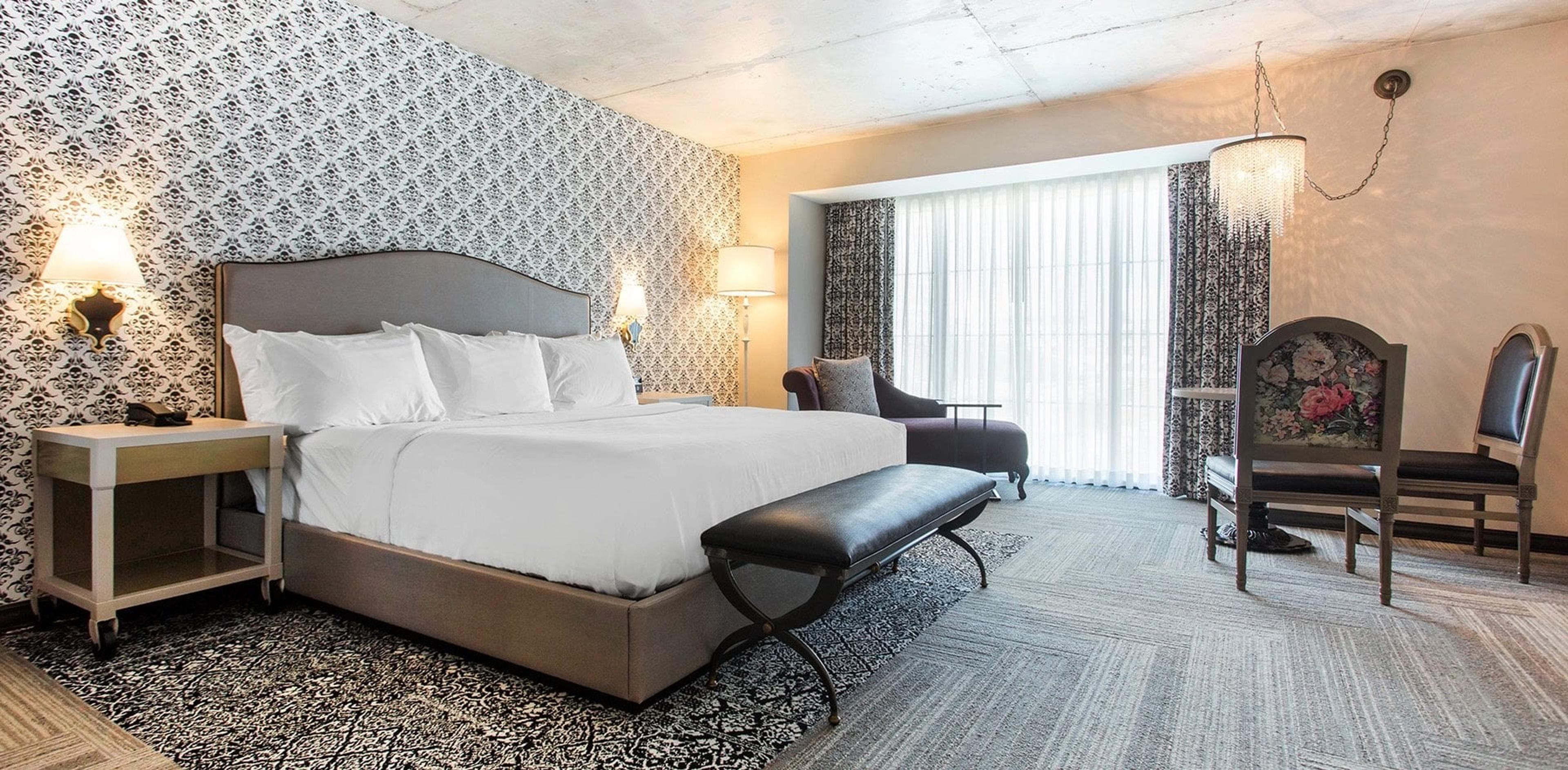 A hotel room with a king bed, black and white patterned wallpaper, a crystal chandelier, and a curtained window with a seating nook beside it.