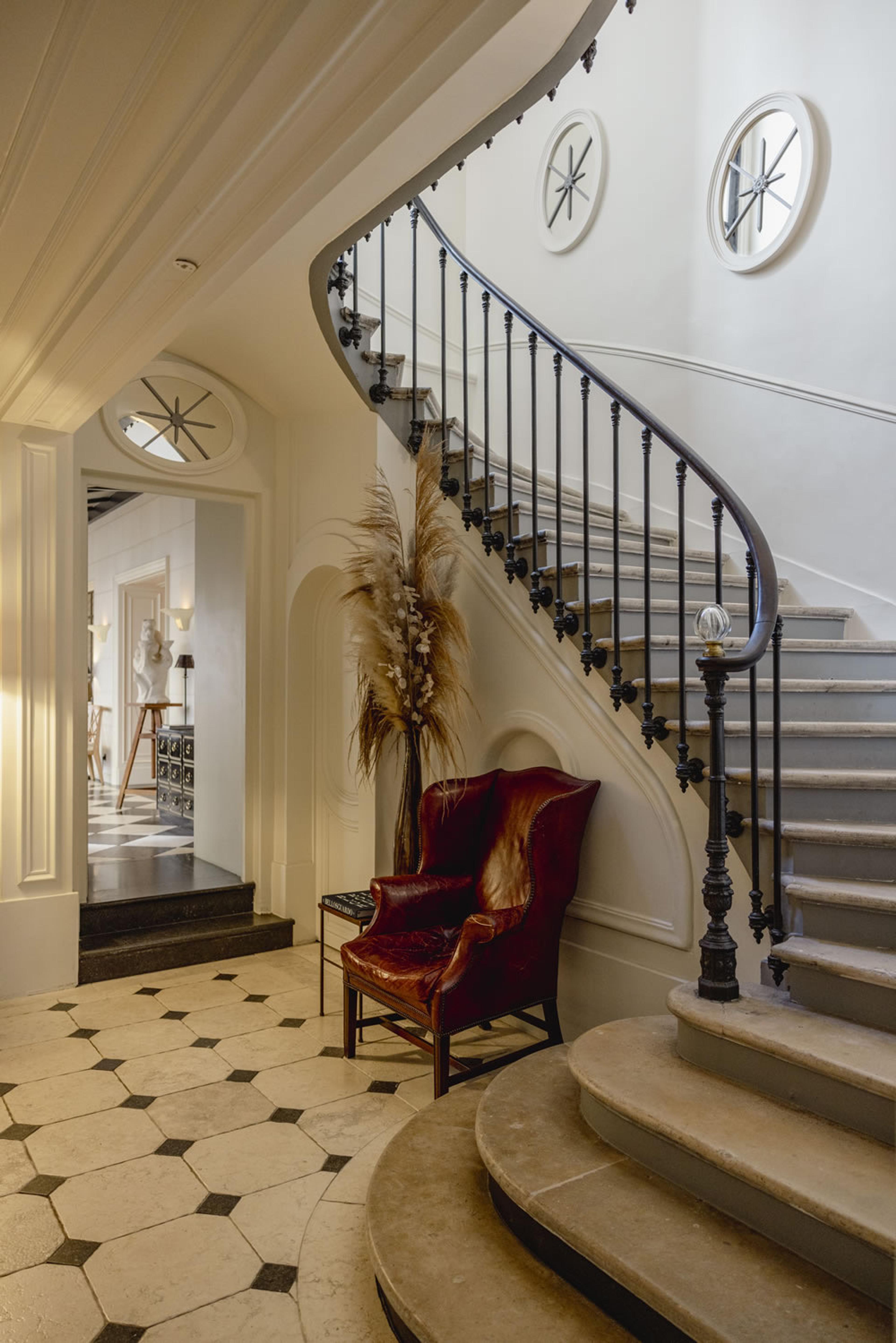 A curved stone staircase with a wrought iron banister, a round window above, and a red leather wingback chair on the black and white tiled landing below.