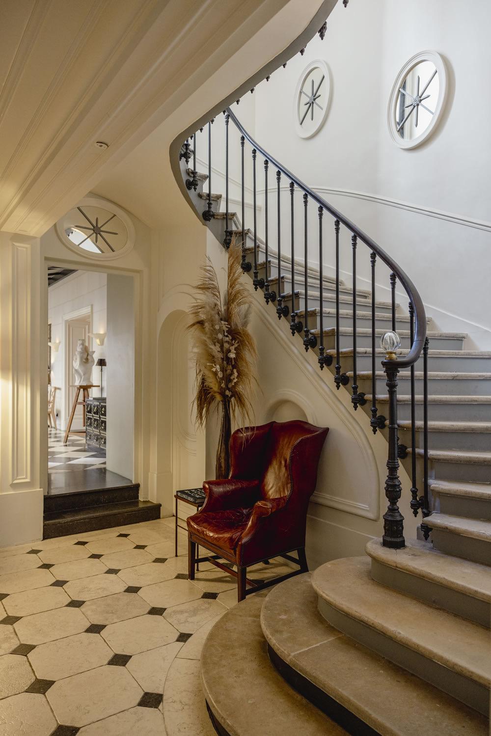 A curved stone staircase with a wrought iron banister, a round window above, and a red leather wingback chair on the black and white tiled landing below.