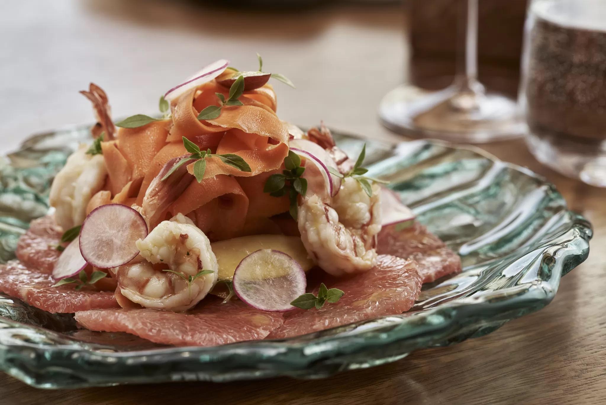 A plate of cured ham, poached prawns, sliced radish and carrot ribbons, with a wine glass out of focus behind it.