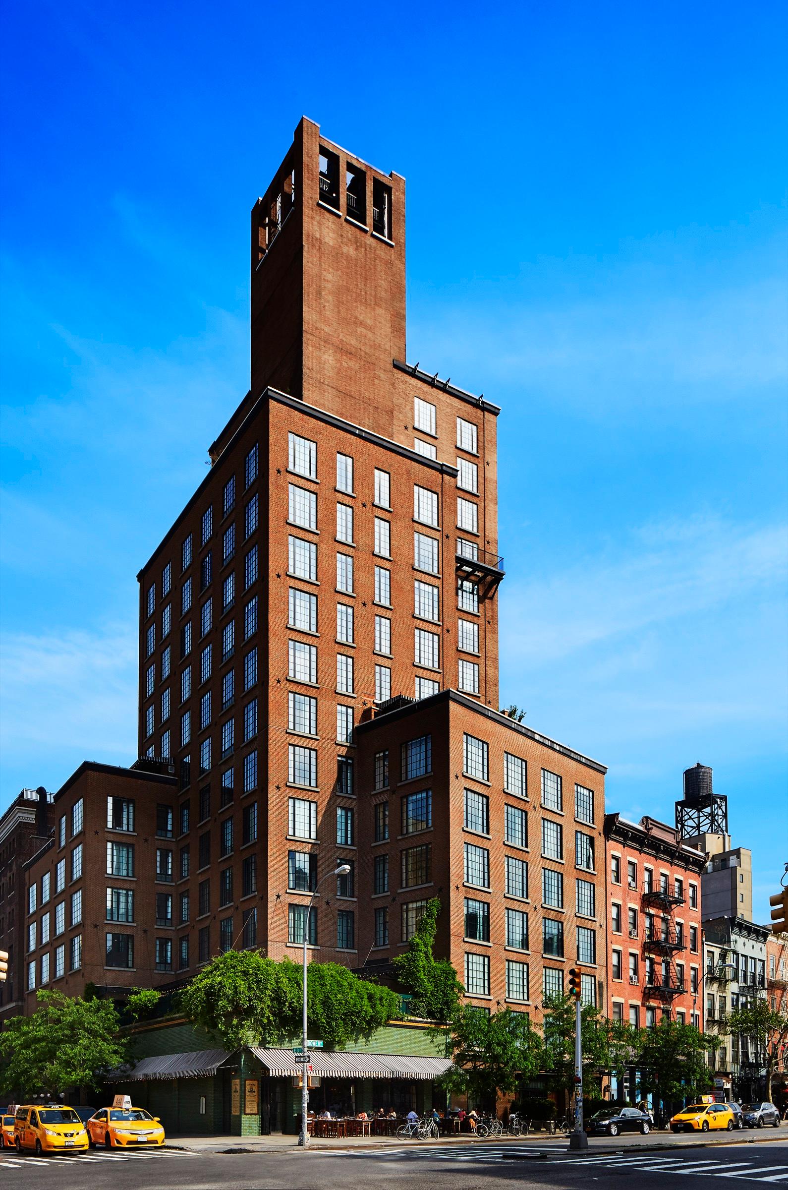 A tall brick hotel tower rises above a Manhattan street corner under a clear blue sky, with yellow cabs passing below.